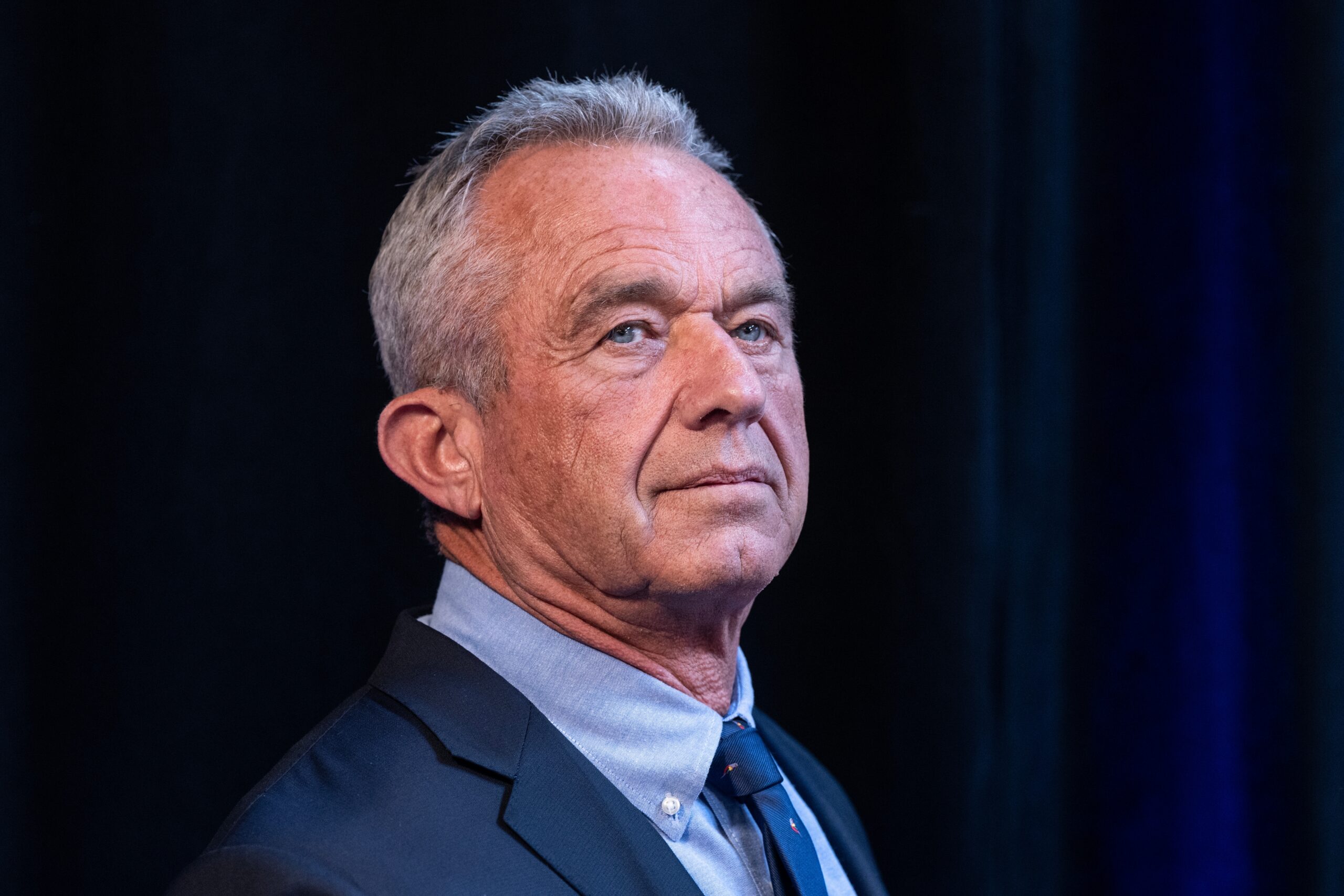 Robert F. Kennedy Jr. looking to the side while wearing a suit and tie against a dark background.