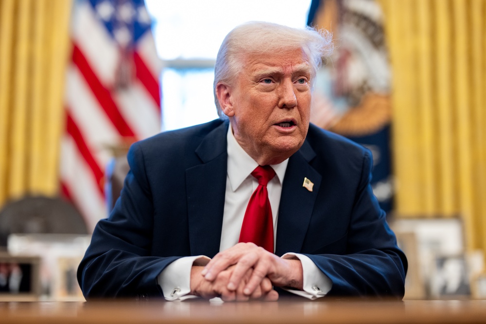 President Donald Trump speaking at desk in Oval Office with American flags behind him.