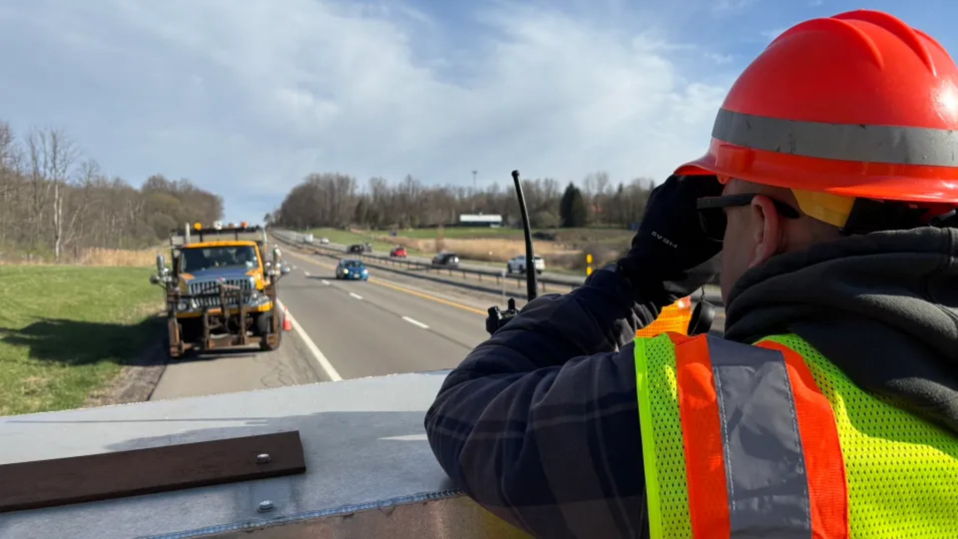An over-the-shoulder shot of a highway construction worker looking through binoculars from an overpass; he wears an orange hard hat, sunglasses, black gloves, and a high-visibility yellow-and-orange safety vest, monitoring traffic and a yellow construction truck on the highway below.