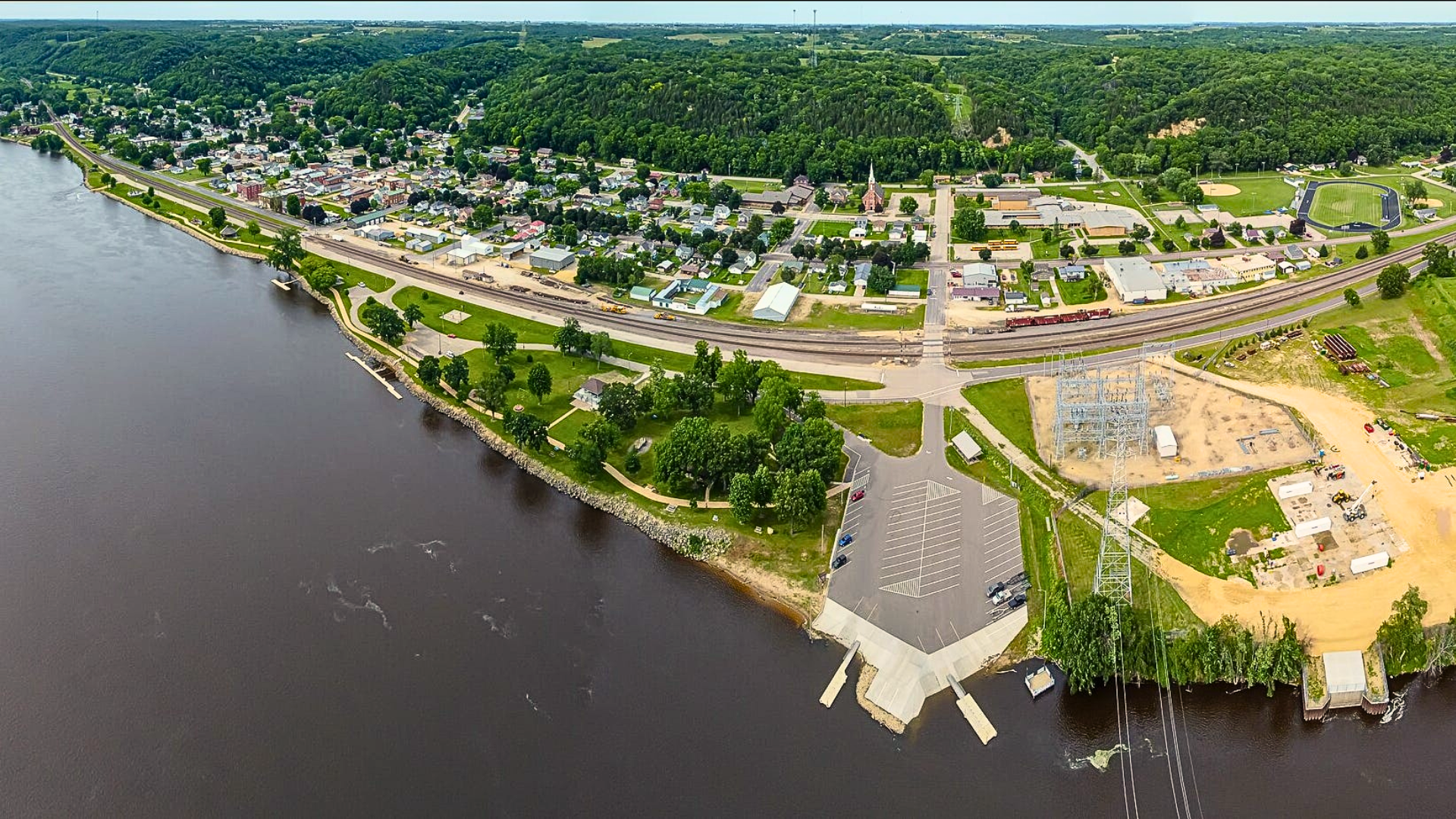An aerial drone shot of the riverfront town of Genoa, Wisconsin, showing rows of houses, a church spire, and a green sports track running alongside a wide Mississippi River and a set of railway tracks.