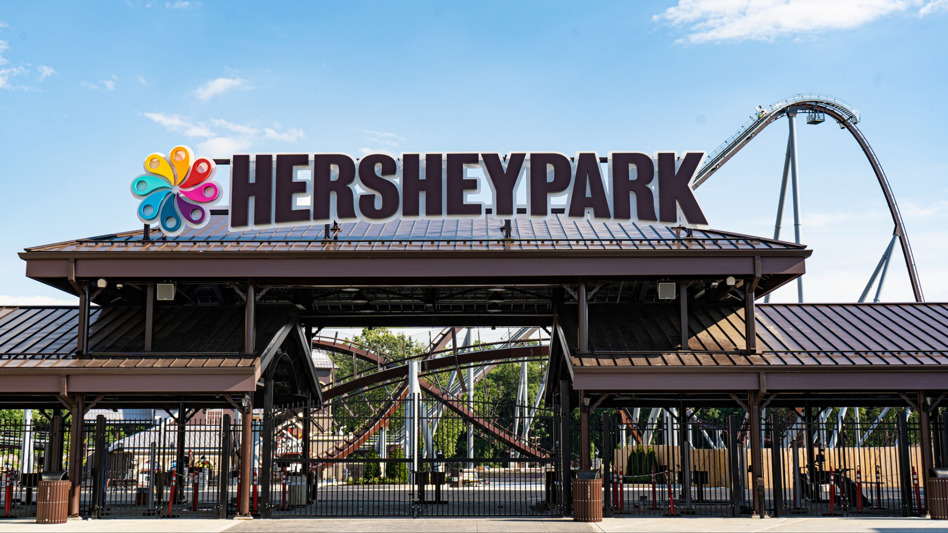 A symmetrical wide shot of the main entrance to Hersheypark on a clear day; a brown metal roof structure supports a massive black sign that reads 
