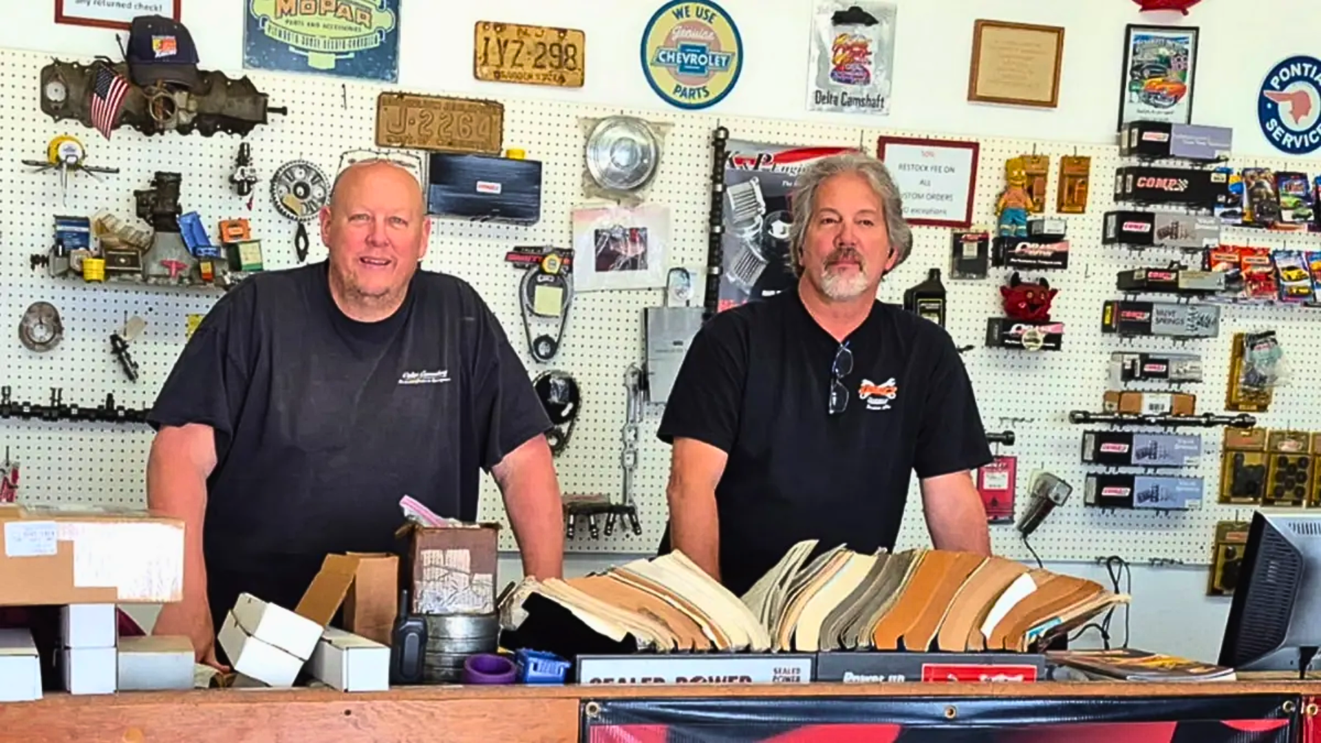 A straight-on indoor shot behind a wooden counter featuring two smiling, middle-aged mechanics in black t-shirts; a pegboard wall behind them is packed with vintage license plates, automotive decals, and packaged engine valves, springs, and camshafts.