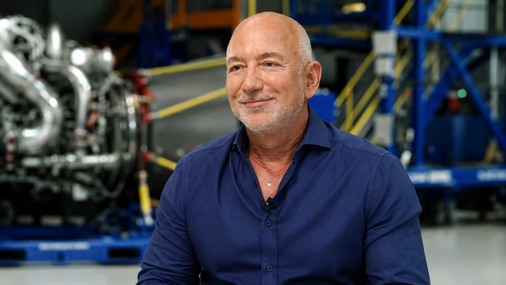 A close-up portrait of Jeff Bezos smiling with a short gray beard wearing a blue button-down shirt, sitting in an industrial facility with heavy machinery blurred in the background.