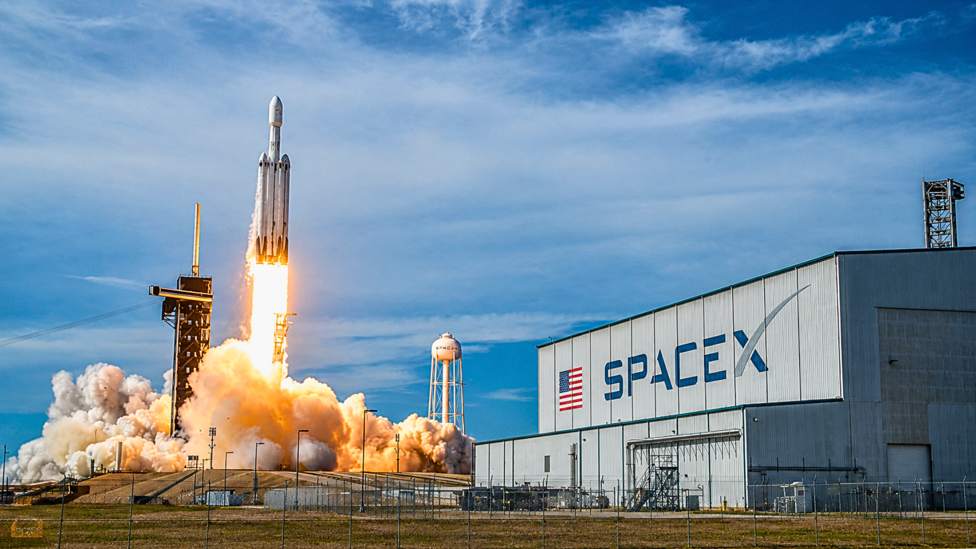 A Falcon Heavy rocket lifts off from the launchpad next to a large white SpaceX hangar building under a bright blue sky.