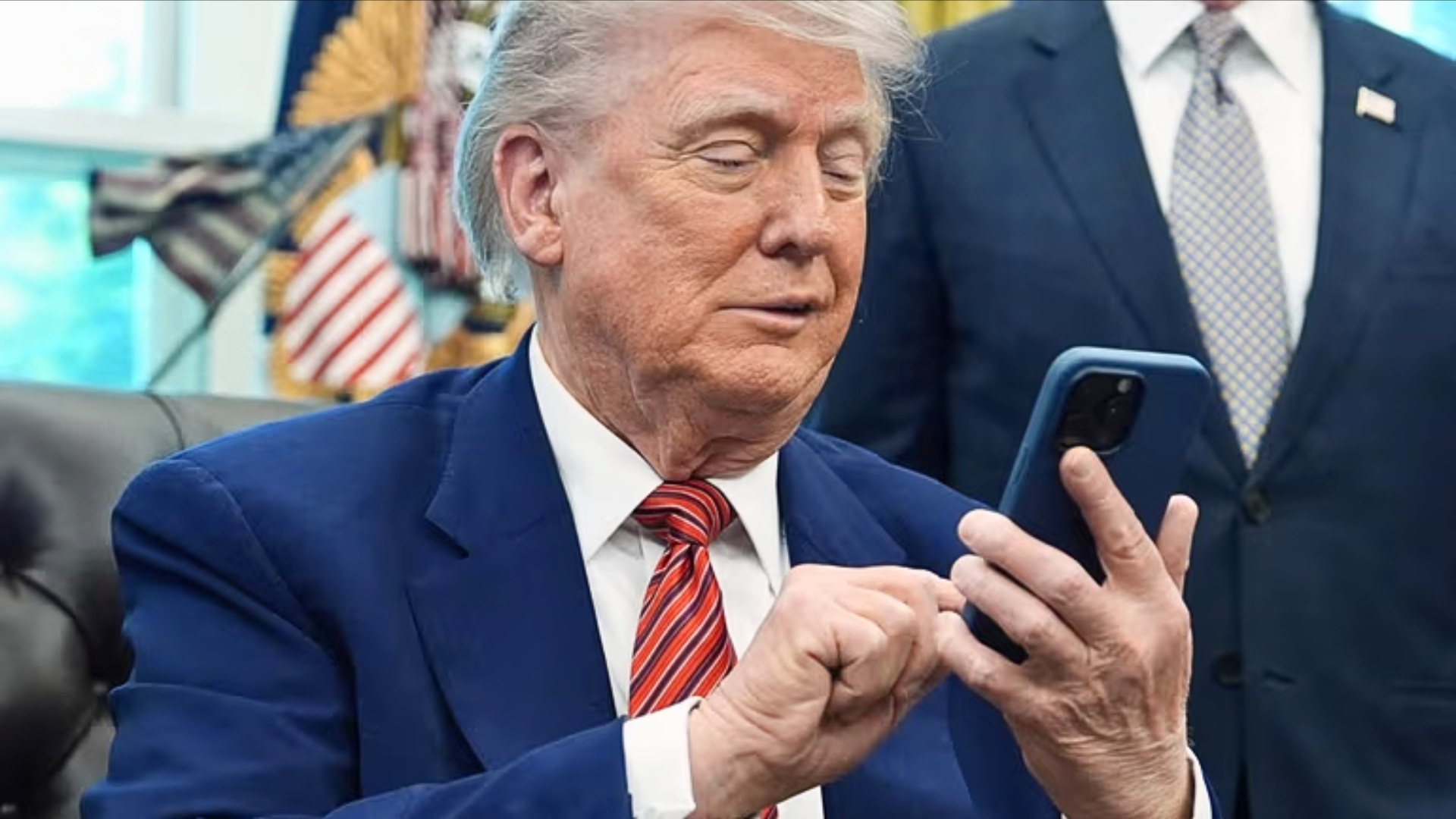 Close-up of Donald Trump in a blue suit and striped red tie, actively scrolling on a smartphone held in a blue case while an assistant stands nearby.