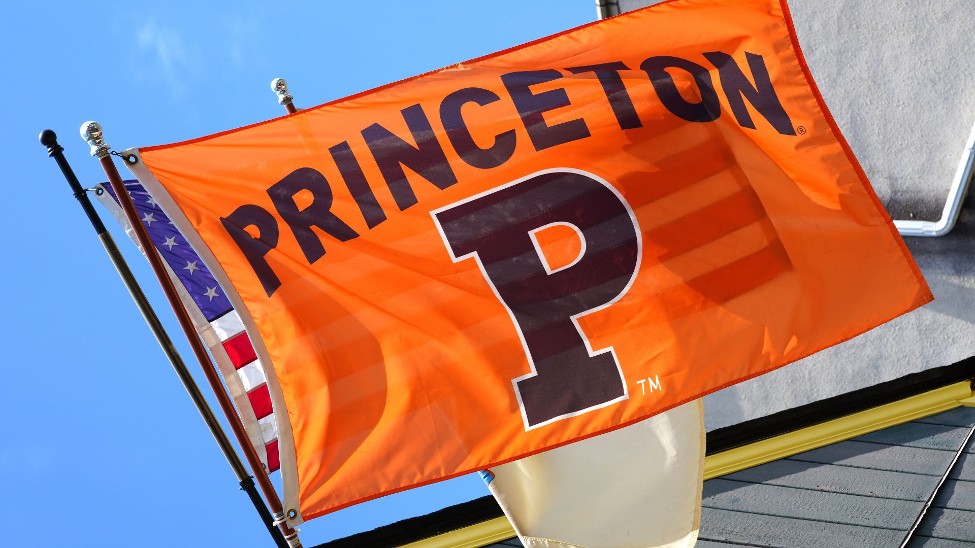 Bright orange Princeton University flag with a large black “P” waves outside beside an American flag under a blue sky.