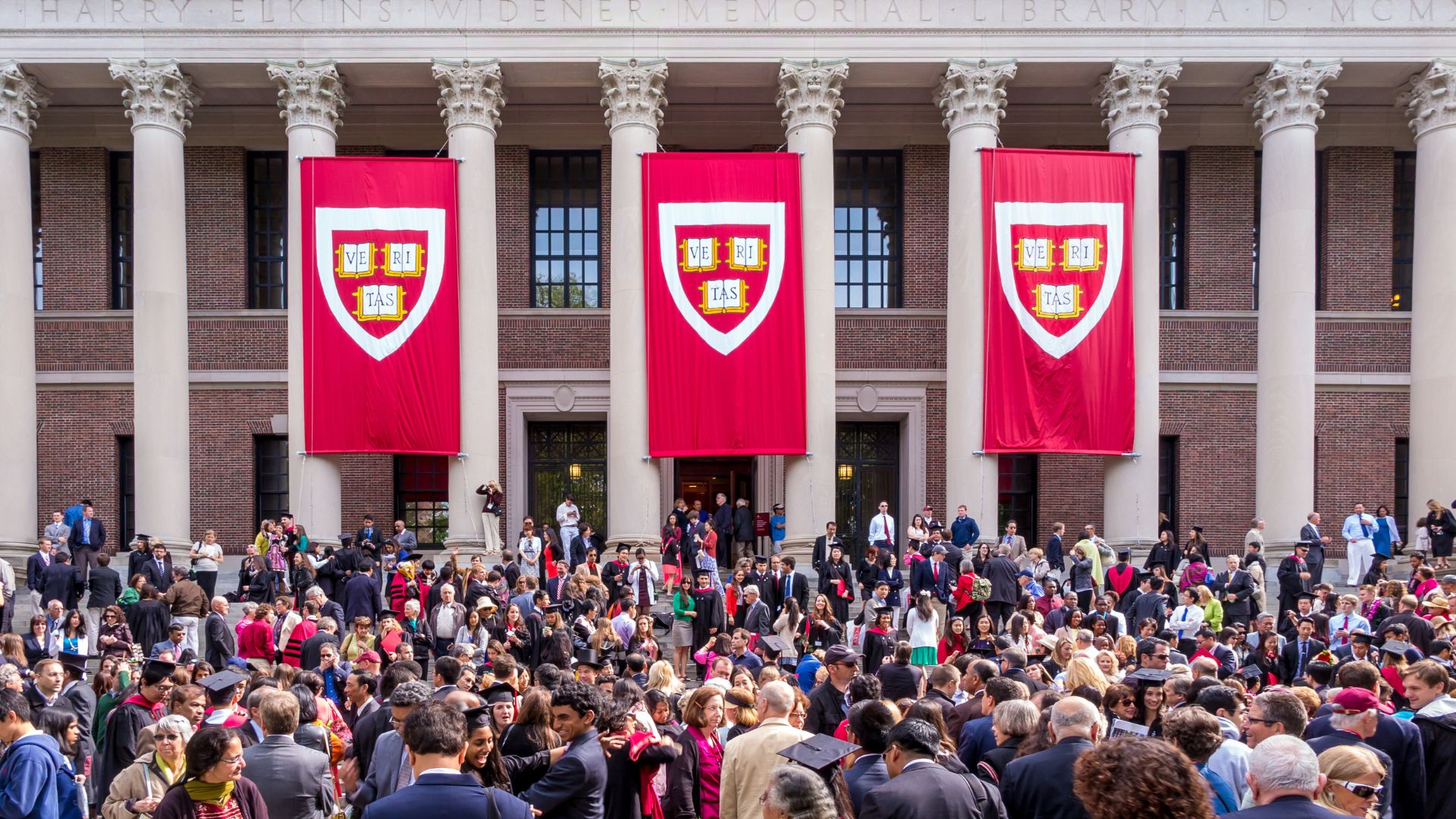 Crowd of students, graduates, faculty, and families gather outside Harvard University's Widener Library beneath three large crimson banners displaying the university crest.