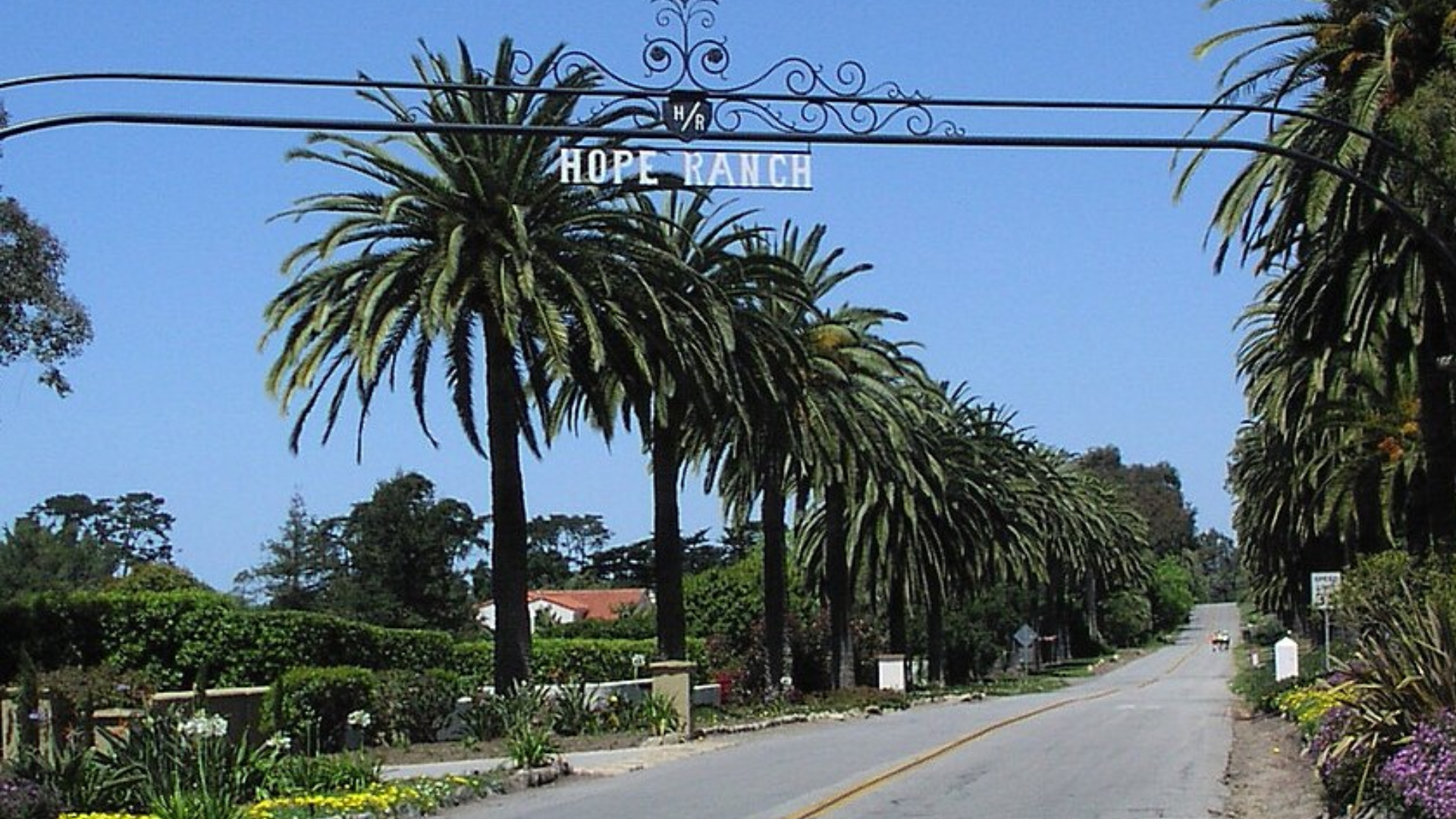 An arched metal sign reading "HOPE RANCH" spans across a paved road lined with tall palm trees.