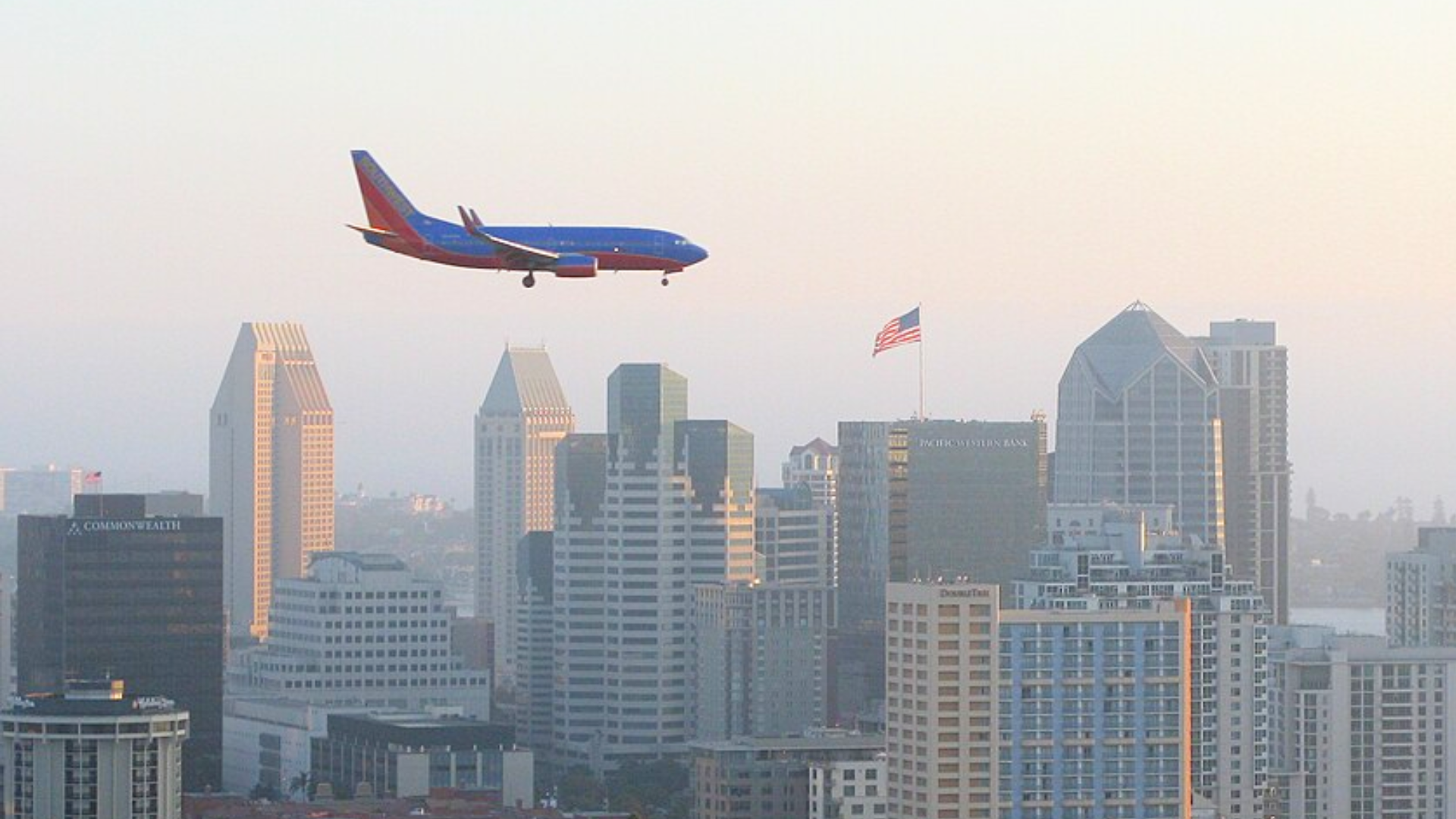 A blue Southwest Airlines plane flies low over a city skyline filled with tall buildings.
