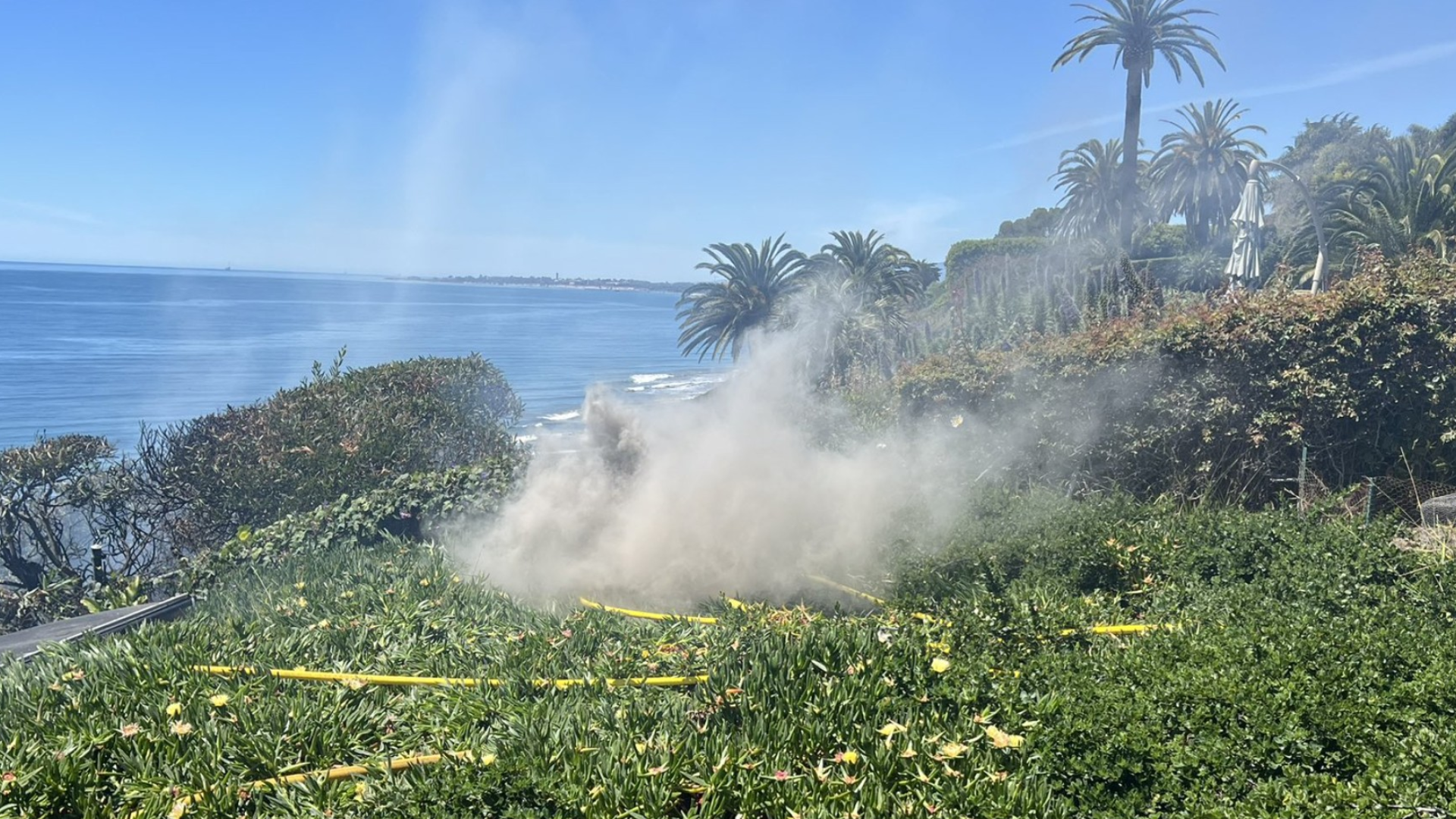 A cloud of white smoke rises from green ground cover on a coastal cliff, with yellow fire hoses snaking through the plants.