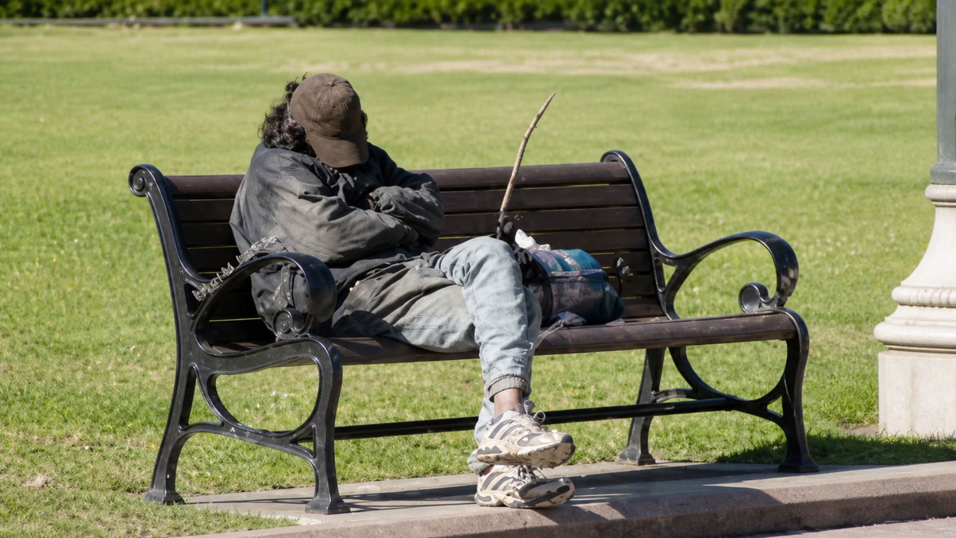 Person sleeping on a park bench with belongings beside them in a grassy public area.