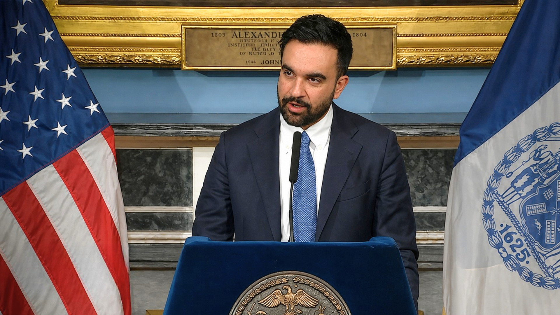 New York City Mayor Eric Adams speaking at a press conference with U.S. and city flags behind him.
