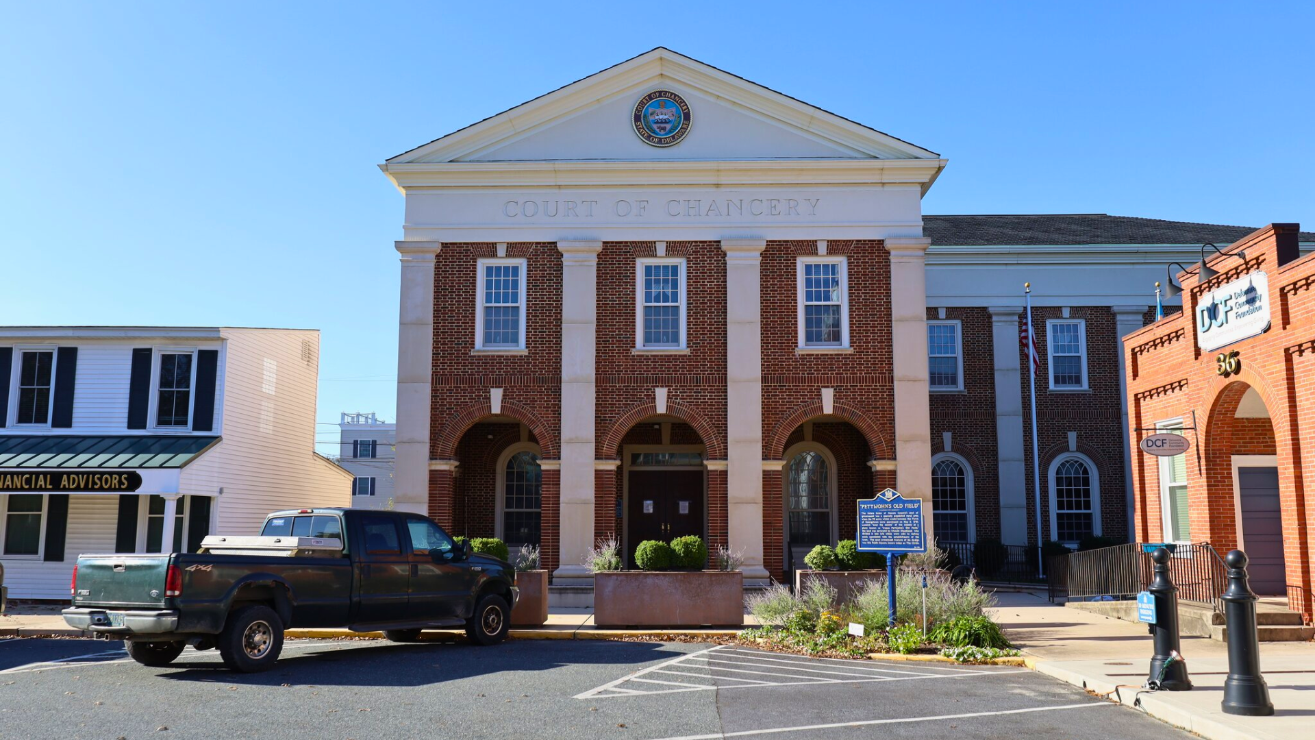 A wide exterior shot of the historic brick Court of Chancery building in Georgetown, Delaware, featuring white columns, arched entryways, and a circular seal on the pediment; a dark green pickup truck is parked in the foreground.