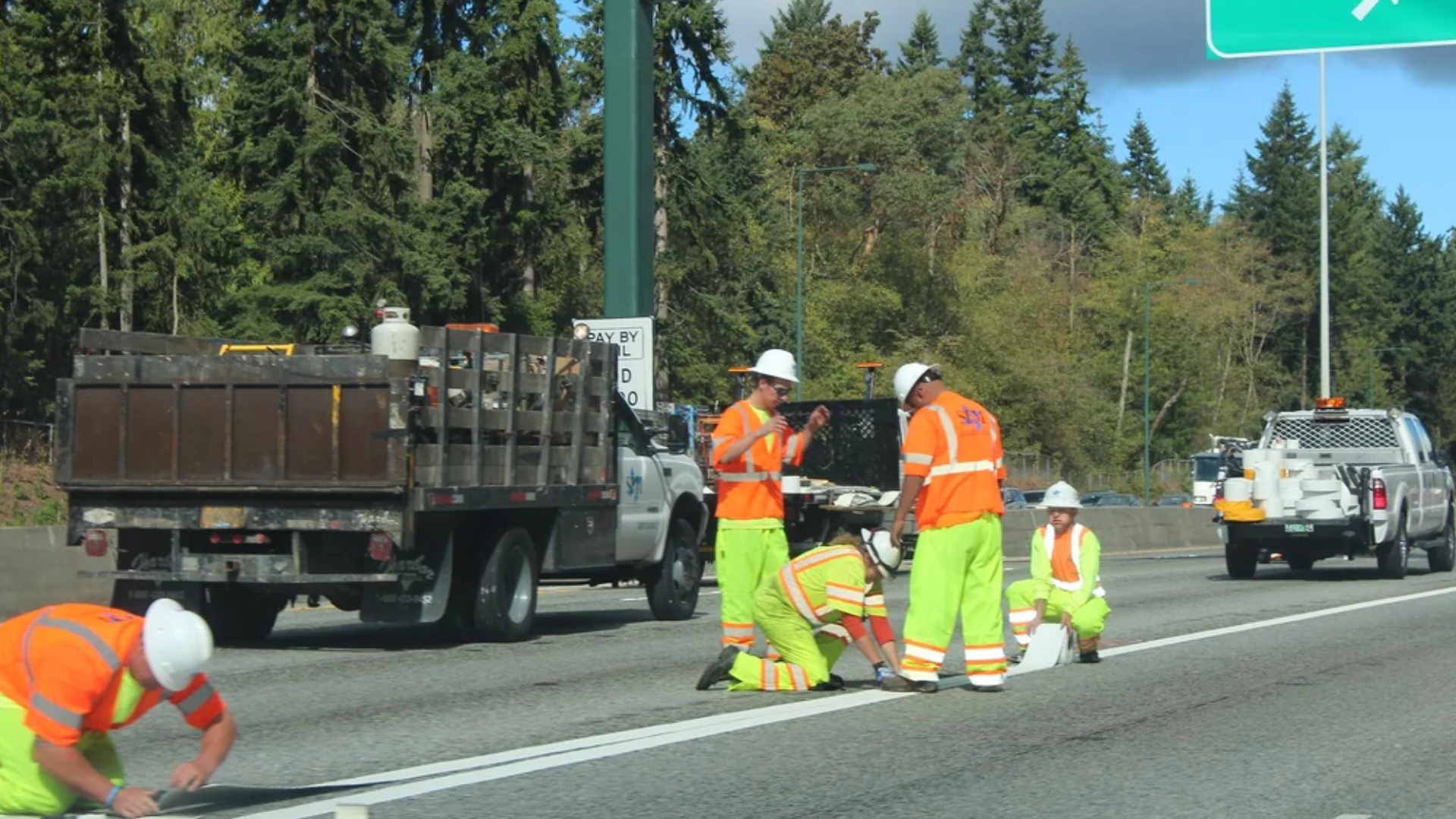 A daytime shot of a highway work zone where several crew members in high-visibility orange jackets and lime-green pants are working together to paint or apply a thick white traffic lane stripe on the asphalt next to a utility work truck.