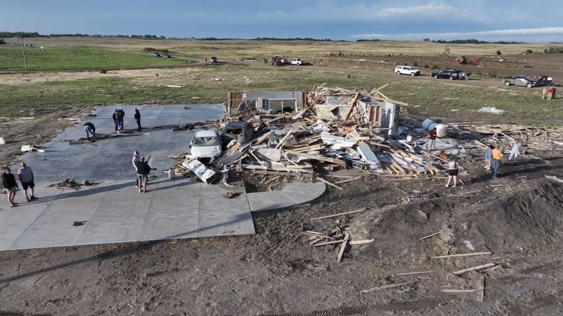 Aerial view of a home completely destroyed by a tornado with debris scattered across a rural landscape as residents inspect the damage.