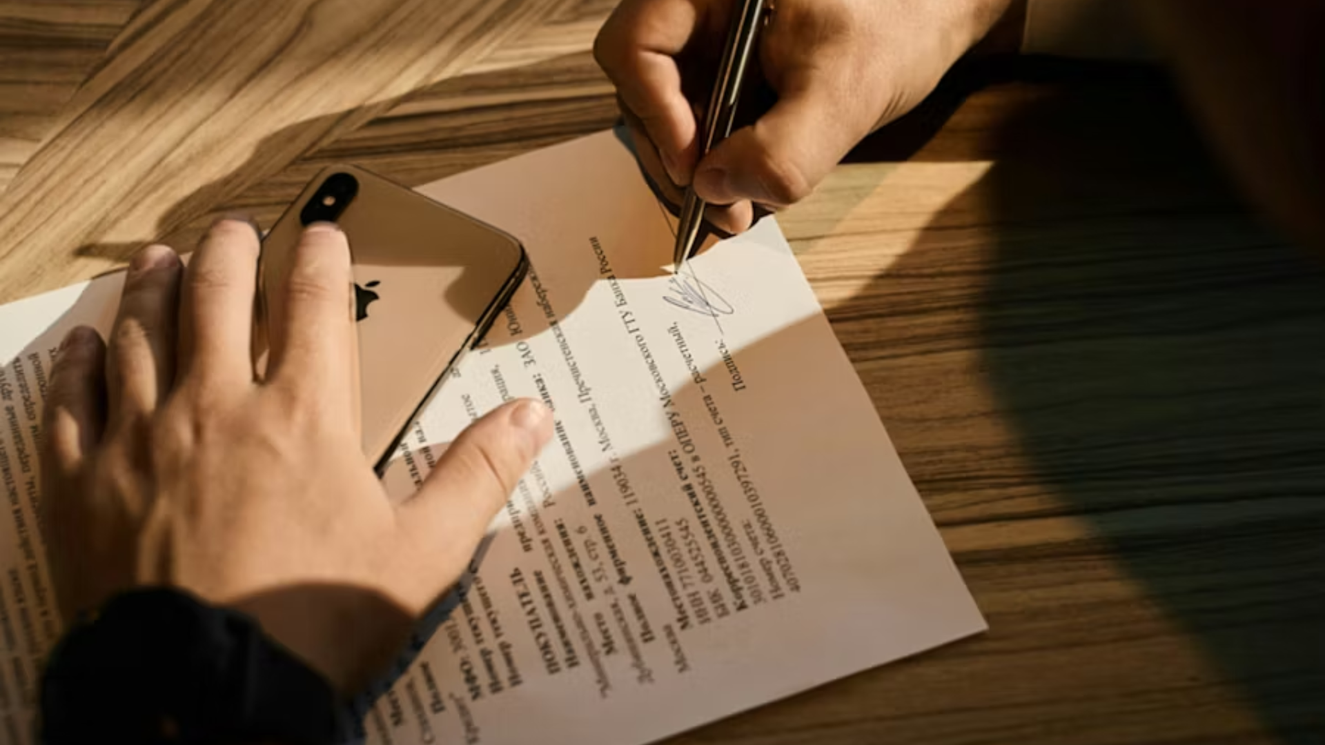 A high-angle close-up of a person's hands signing a document with a silver pen on a light wood desk.