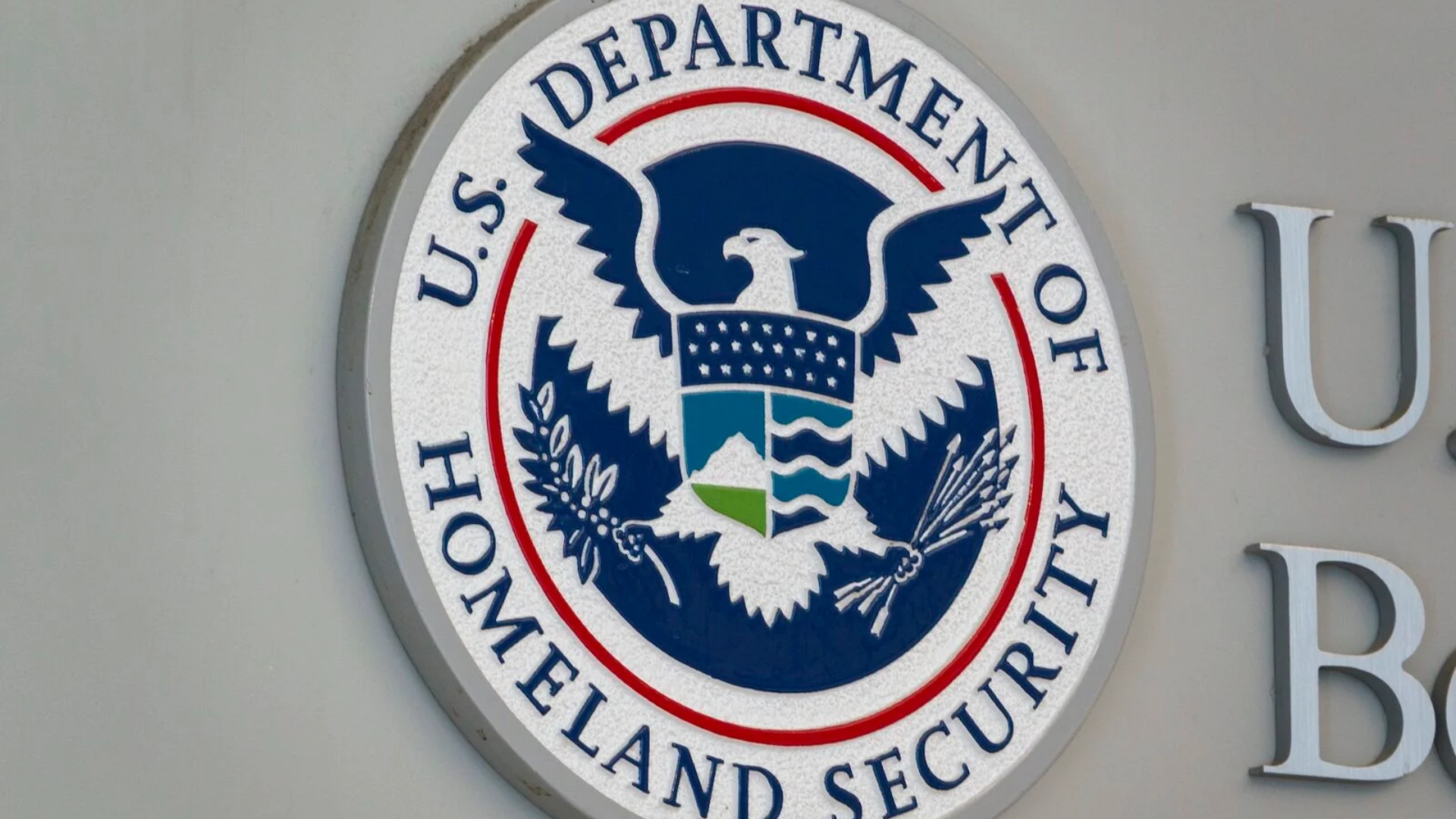 A close-up, sharp-focus shot of the official, circular seal of the U.S. Department of Homeland Security mounted on a light gray office exterior; the emblem displays a white bald eagle with an American shield over a blue background encircled by the agency's name.