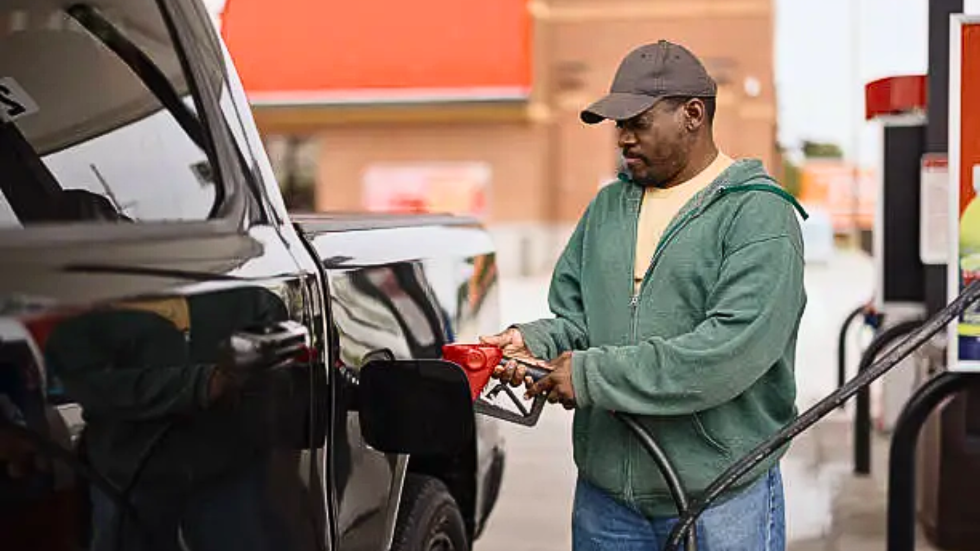 A medium shot of a man wearing a baseball cap and a green hoodie standing at a gas station pump, inserting a fuel nozzle into the tank of a black pickup truck.