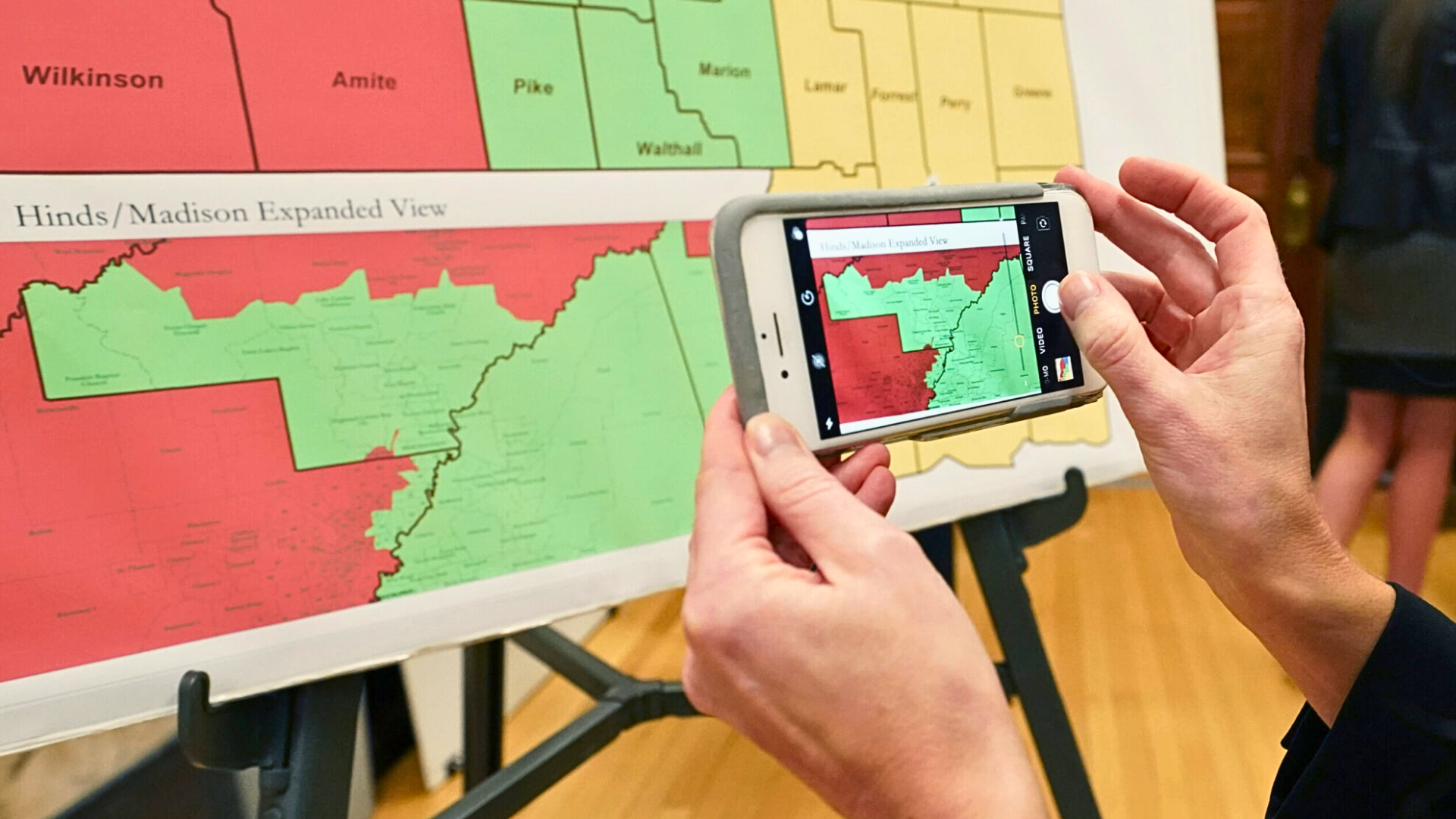 A close-up shot of hands using a smartphone to take a photo of a color-coded "Hinds/Madison Expanded View" redistricting map on an easel.