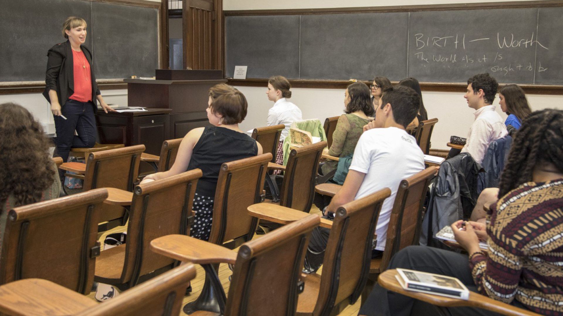College students sit in wooden lecture hall seats while a professor speaks at the front of a classroom with chalkboards behind her.
