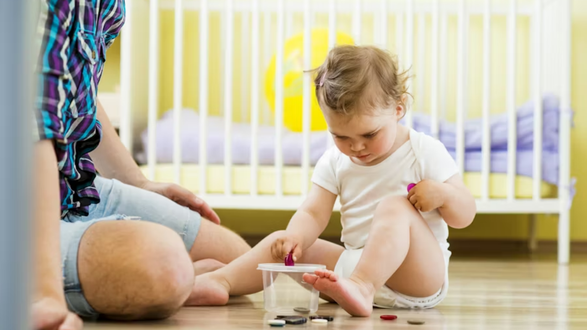 A high-angle shot of a young toddler in a white onesie sitting on a light wood floor, focused on placing small purple objects into a clear plastic container; a man in a plaid shirt and denim shorts sits nearby, supervising the child's developmental play.