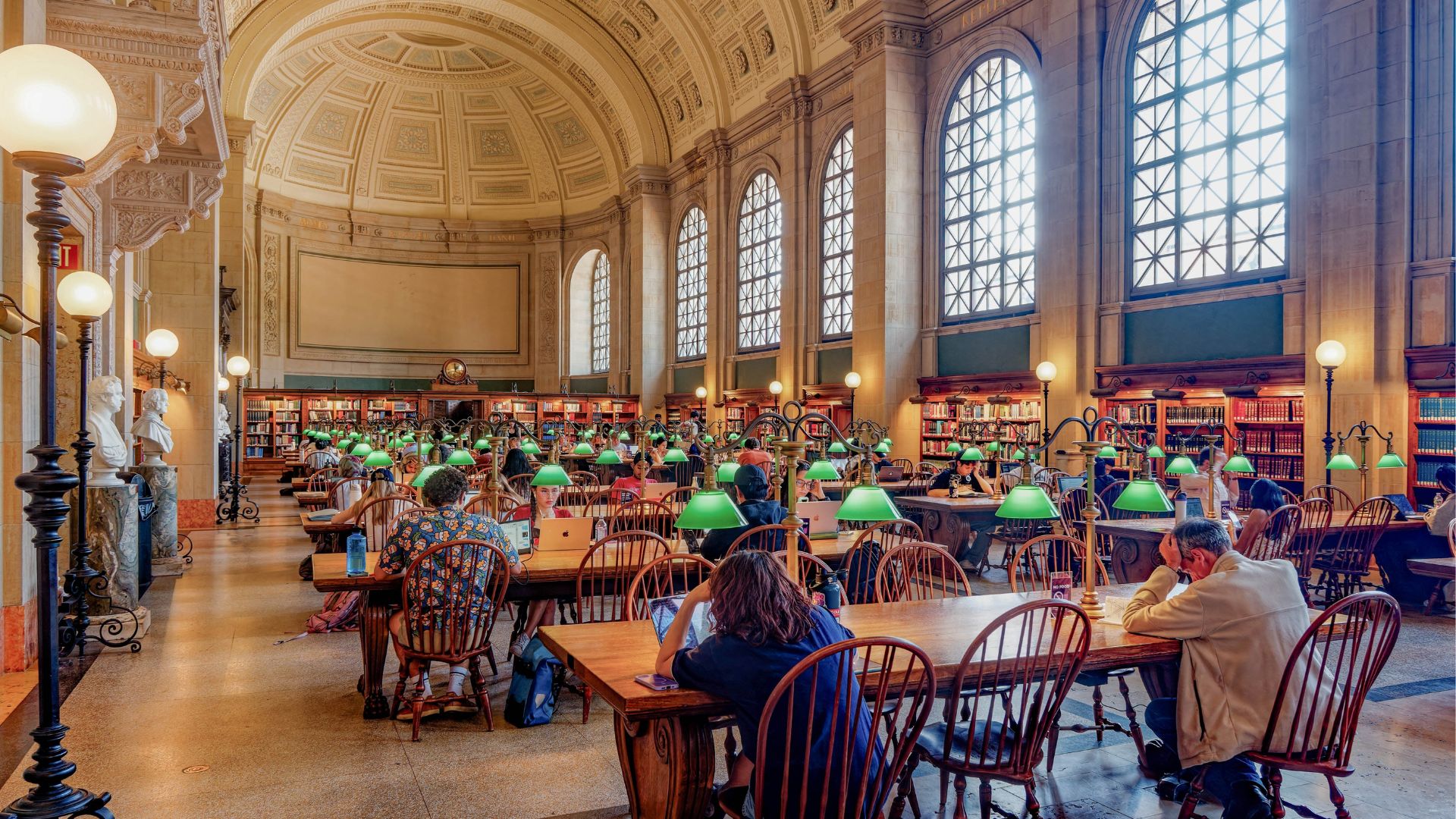 Students study at long wooden tables inside a grand Harvard library reading room with arched windows, vaulted ceilings, and green reading lamps.