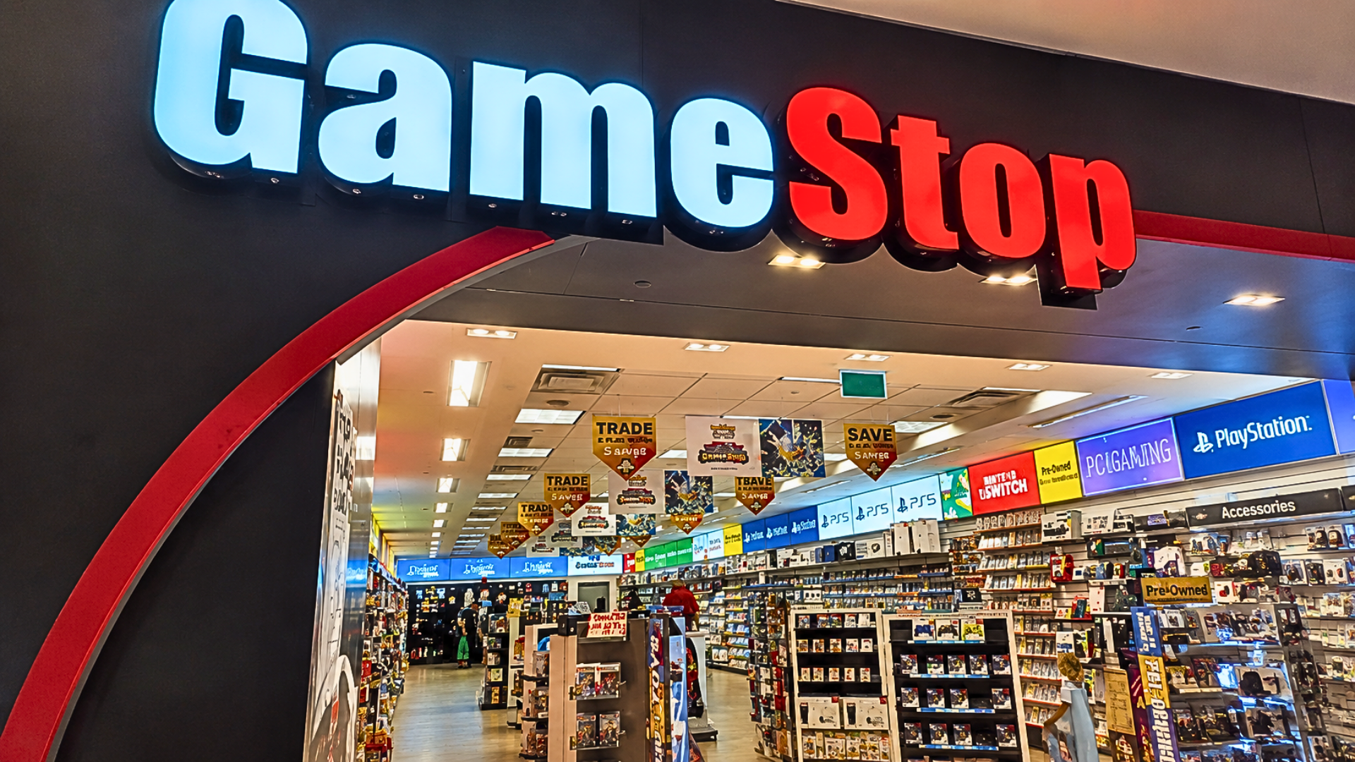 A wide daytime shot of a GameStop store entrance in a brightly lit shopping mall; the iconic blue-and-white neon "GameStop" logo is prominent above the glass doors, which reveal interior shelves filled with Nintendo, PlayStation, and Xbox merchandise and "Trade-In" banners.