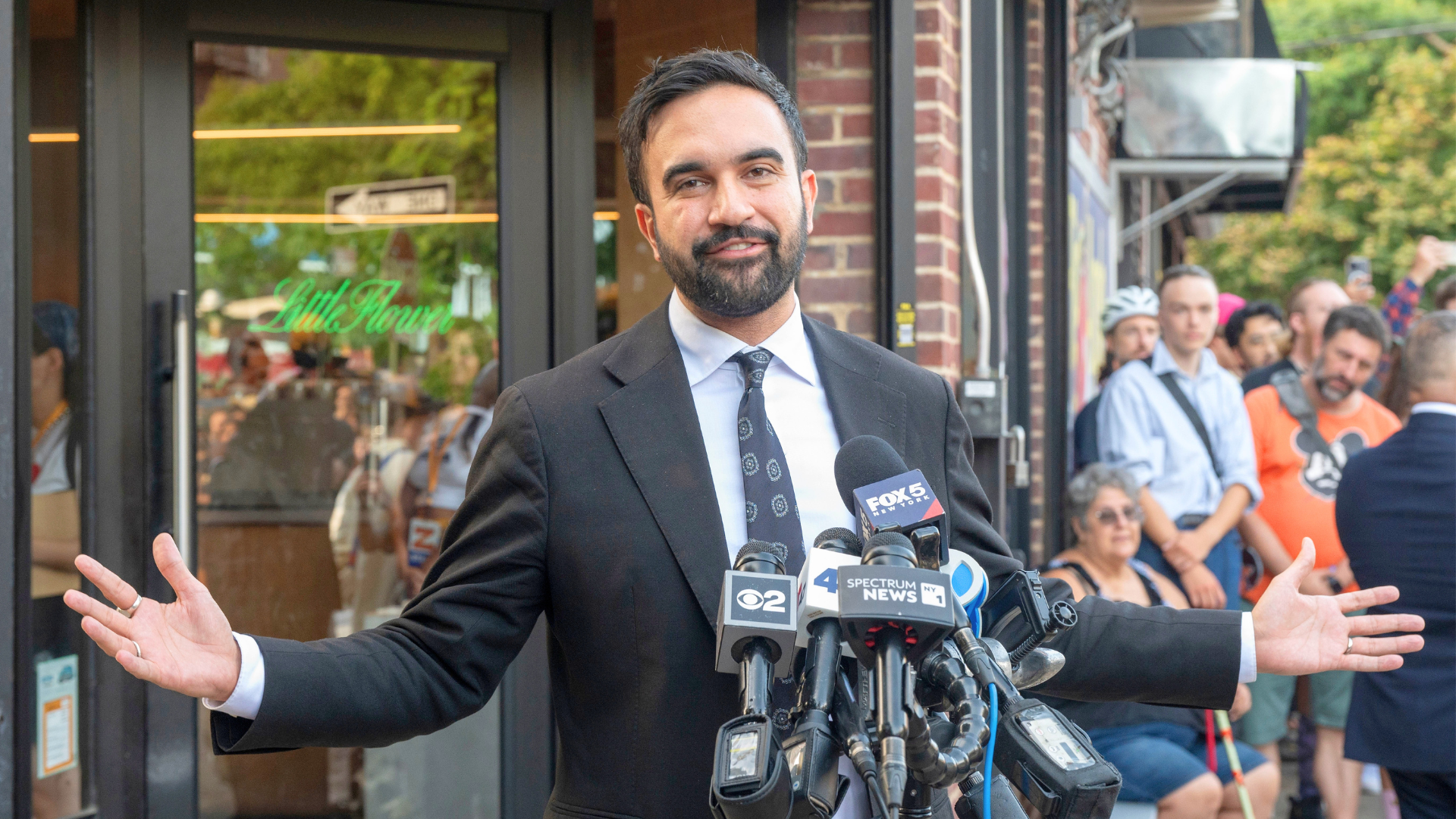 Zohran Mamdani gestures with his hands while speaking to a cluster of news microphones on a city street.
