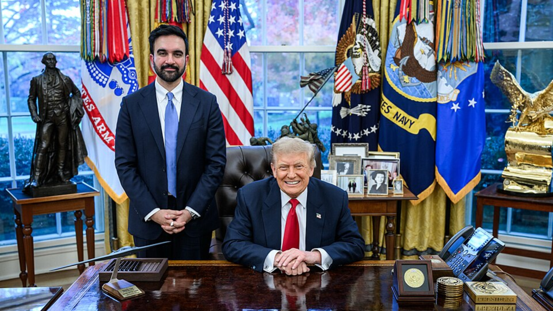Zohran Mamdani stands beside a smiling Donald Trump seated at the Resolute Desk in the Oval Office.