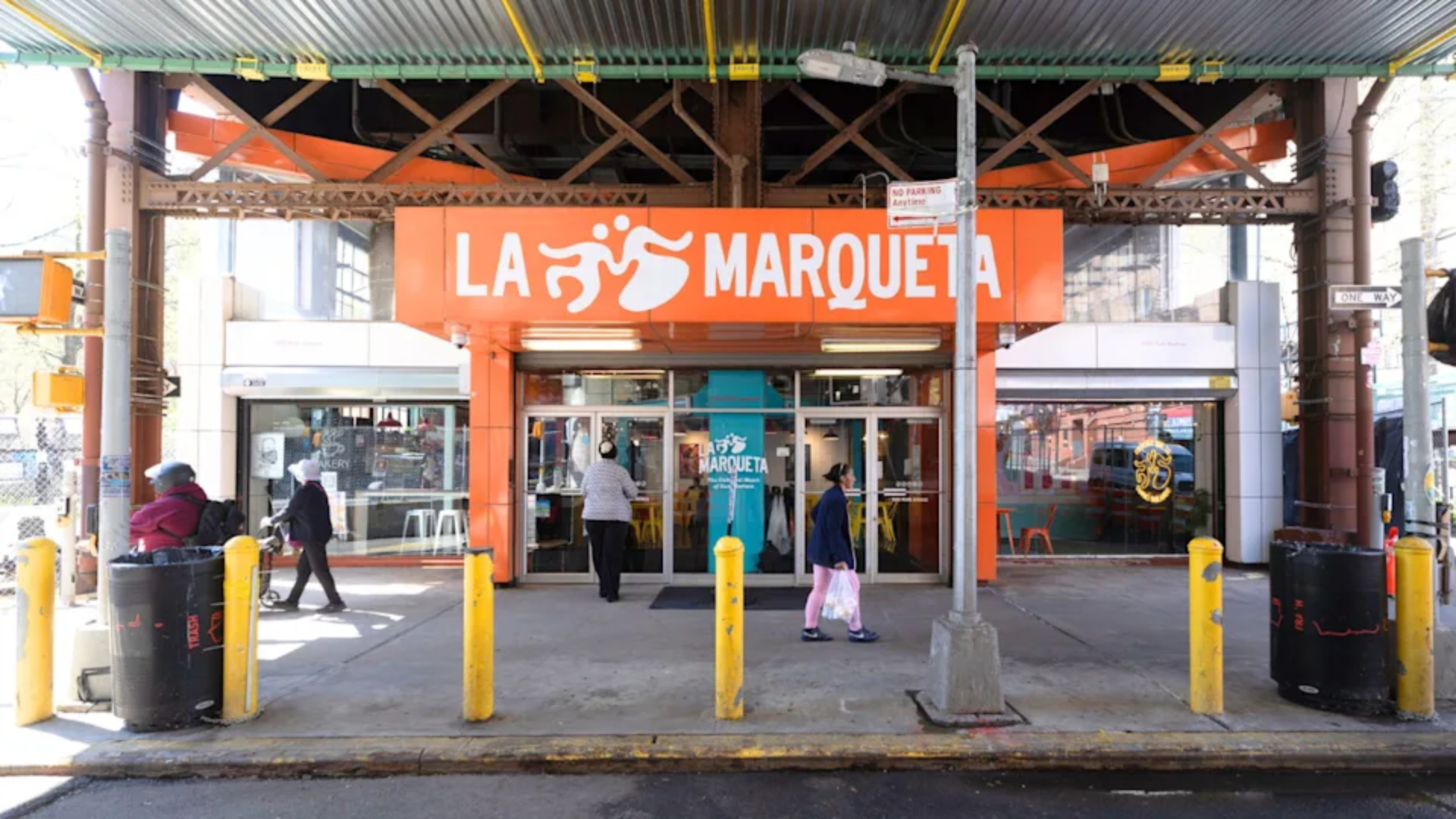 Pedestrians walk past the bright orange storefront of "LA MARQUETA" located under an elevated train structure.