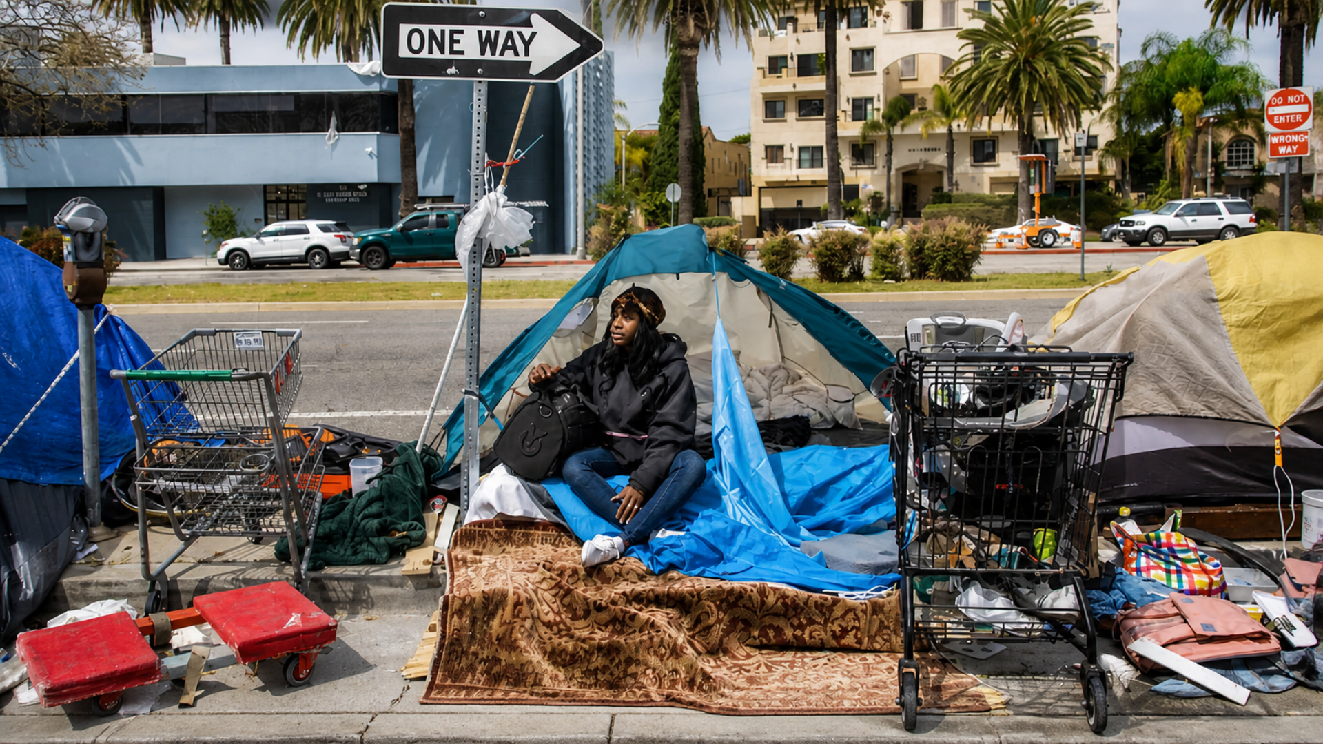 Woman sitting in a sidewalk tent surrounded by belongings and shopping carts along a city street.