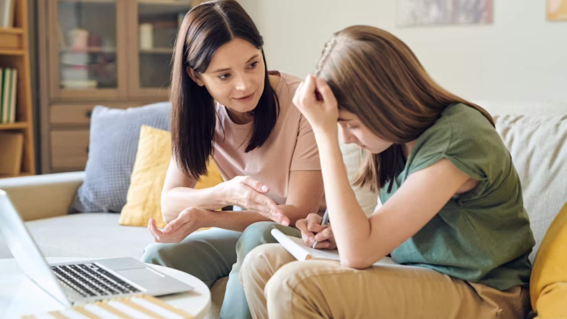 A medium shot of a concerned mother on a sofa talking softly to her teenage daughter, who looks stressed and rubs her temple while trying to write in a notebook.