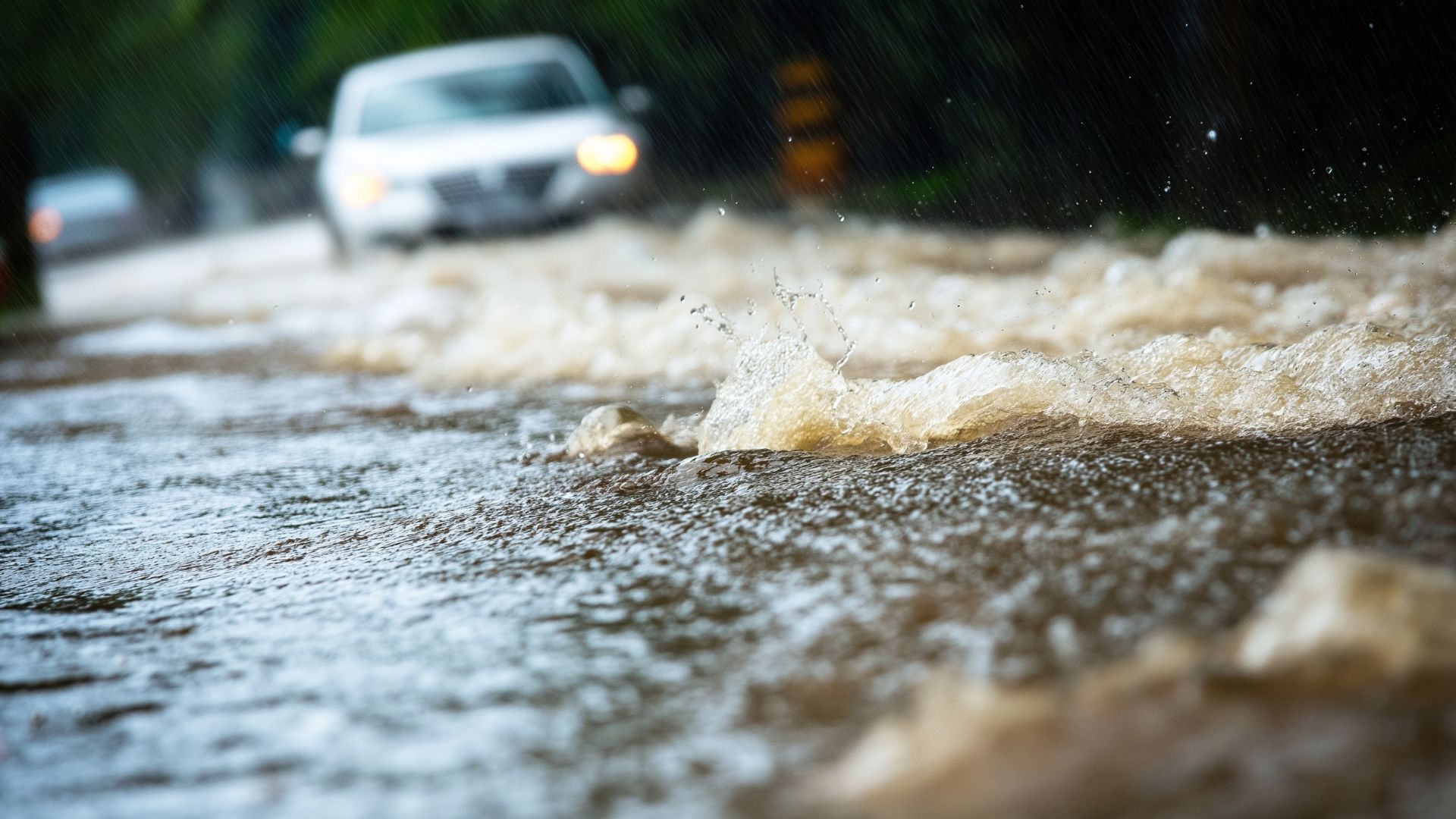 Floodwater rushes over a roadway while cars drive through heavy rain with headlights on.