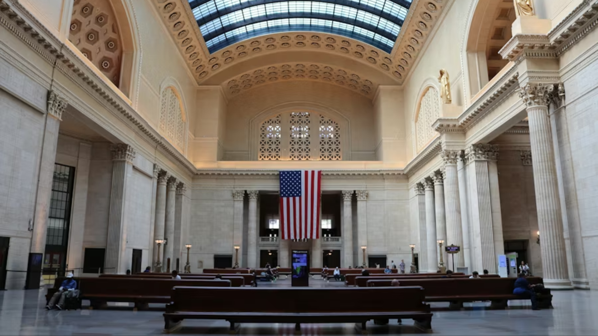 A wide interior shot of the historic Chicago Union Station Great Hall; the massive vaulted ceiling and skylight tower over rows of empty wooden benches, with an American flag hanging at the far end, currently serving as a focal point for May Day labor gatherings.