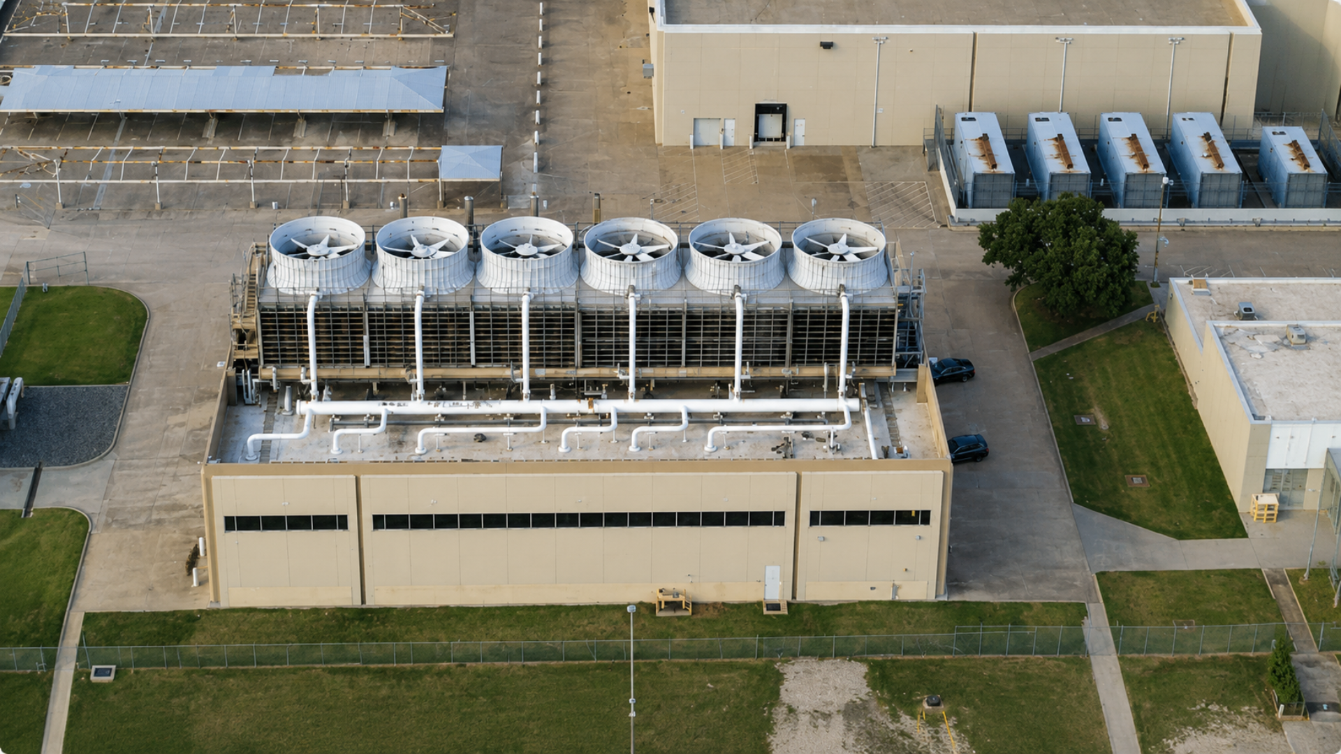 A high-angle aerial view of a massive, tan industrial utility facility featuring six large, white evaporative cooling tower fans arranged in a row with white piping network over a concrete lot.