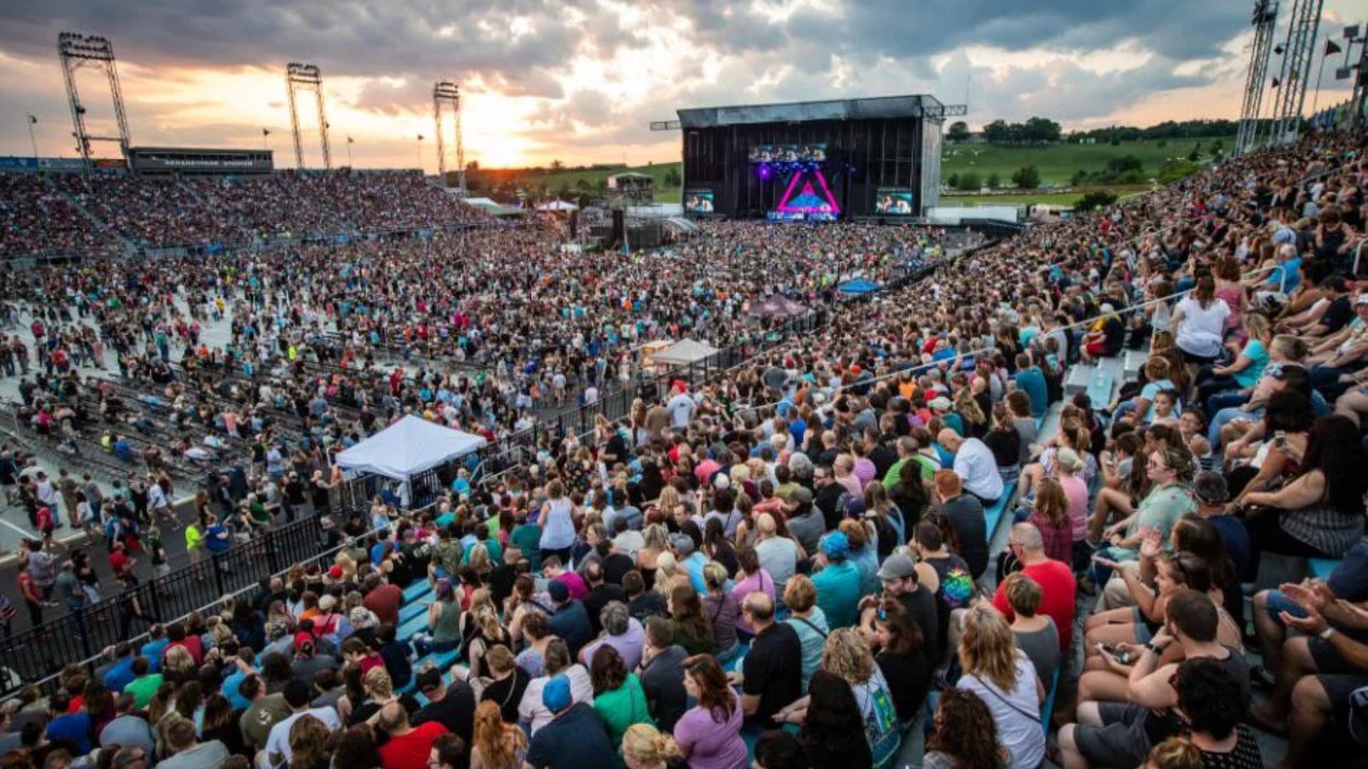 A panoramic high-angle view of a massive concert crowd filling Hersheypark Stadium at sunset; tens of thousands of spectators fill the lower field and the sweeping open-air grandstands under a dramatic cloudy sky as the stage lights glow in the distance.
