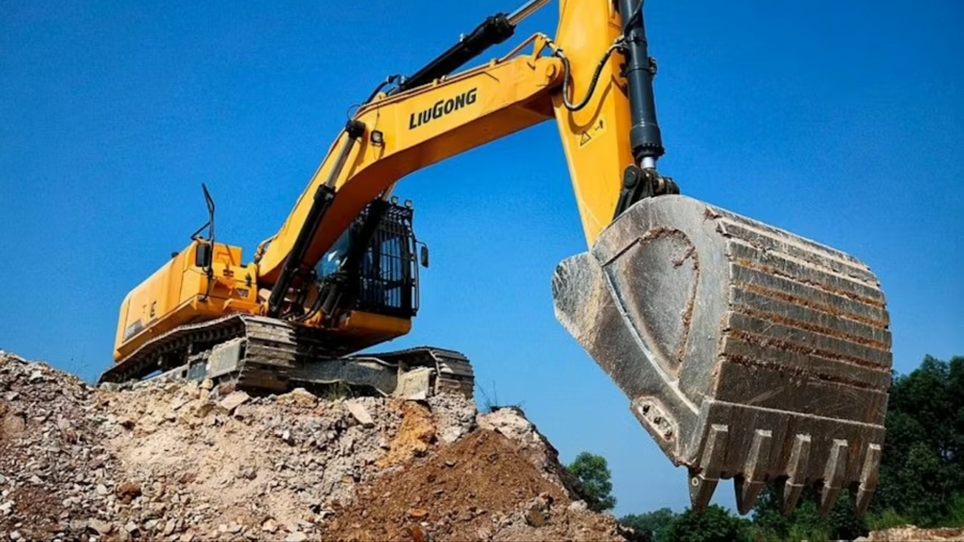 A wide daytime shot of a large yellow LiuGong hydraulic track excavator perched atop a pile of crushed gravel and dirt; its heavy steel digging bucket is extended forward under a clear, bright blue sky.