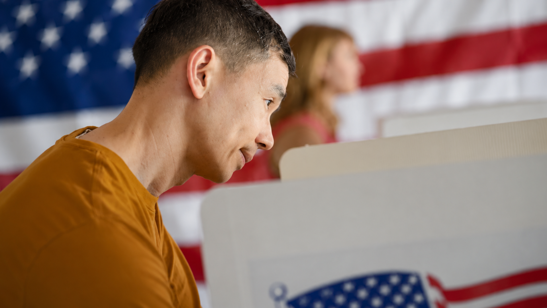 A shallow-focus side profile shot of a young man inside a private cardboard voting booth, looking down intently as he fills out his paper ballot; a blurred American flag hangs in the background behind him.
