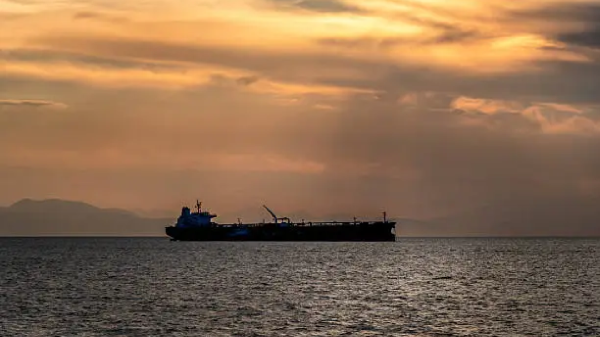 A wide landscape shot displaying the silhouette of a massive cargo ship or oil tanker floating on calm ocean water under a hazy, golden sunset sky.