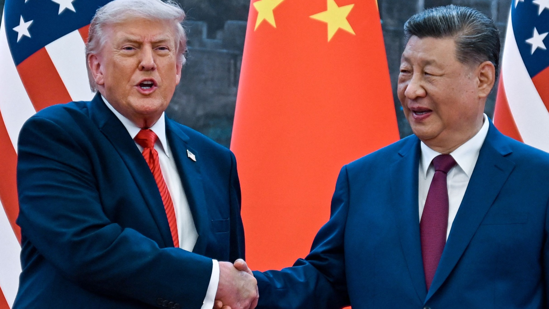 A medium shot of Donald Trump and Chinese President Xi Jinping shaking hands, positioned in front of a backdrop featuring both American and Chinese national flags.