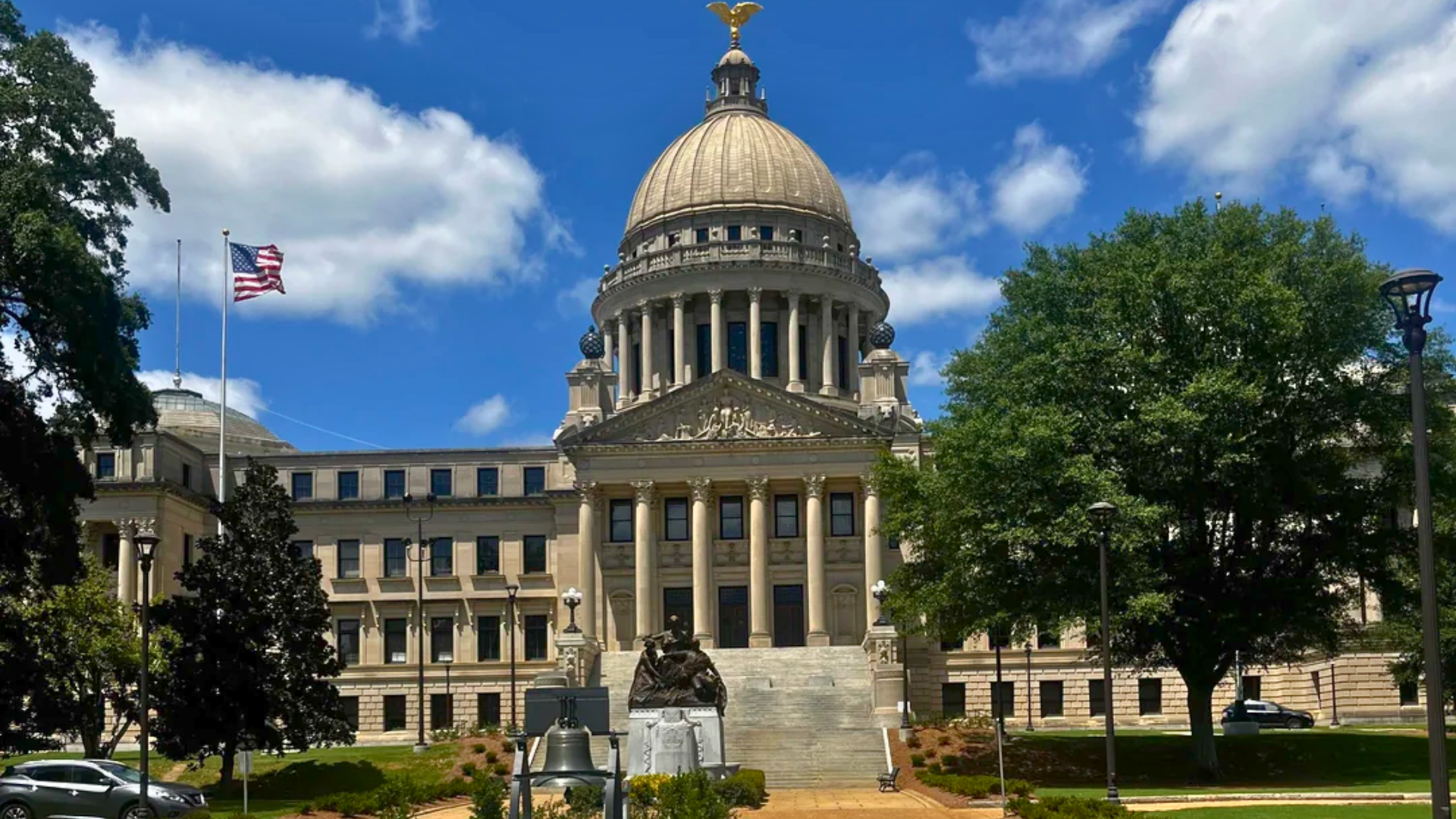A wide exterior view of the Mississippi State Capitol building featuring its grand central dome, gold eagle statue, and classical columns under a blue sky.