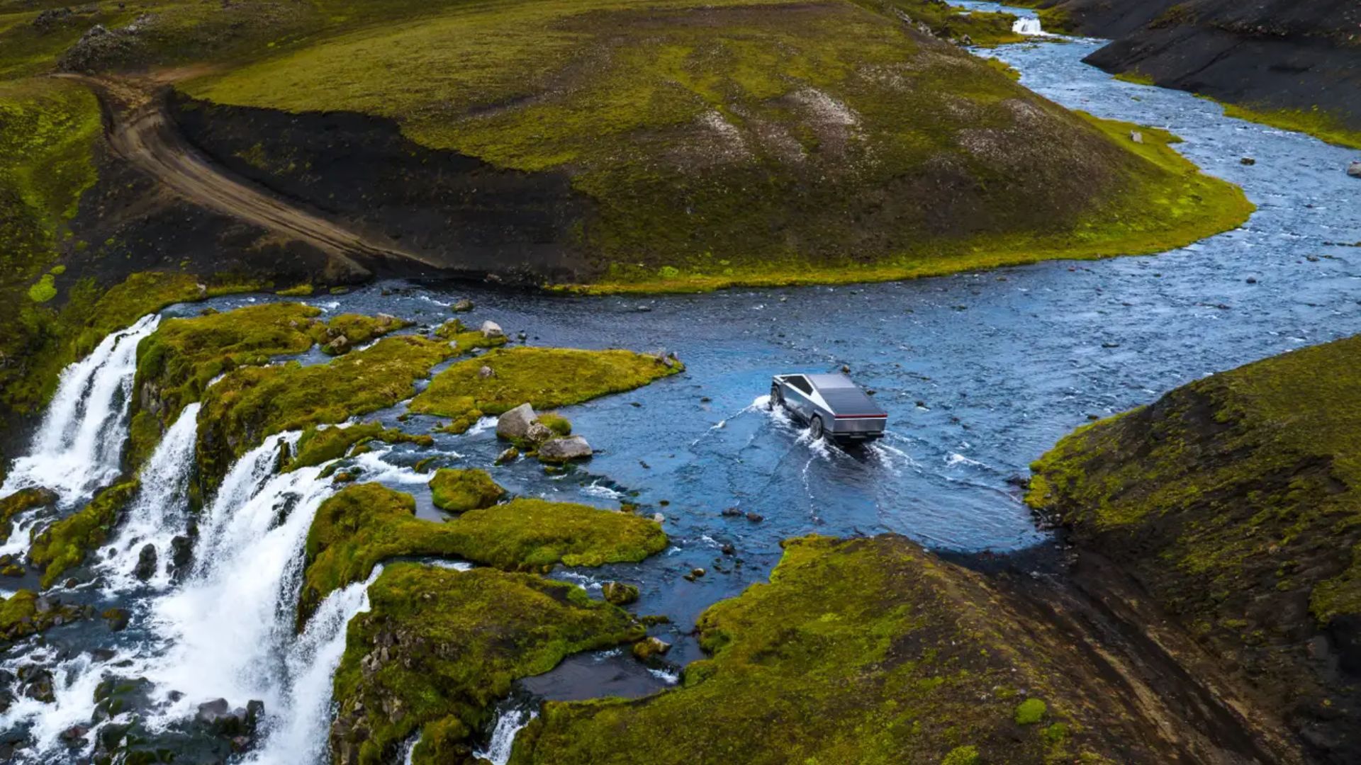 Tesla Cybertruck driving through a shallow river in a remote green landscape with waterfalls and rocky hills surrounding the water crossing. The scene appears to promote the truck’s off road and water driving capabilities tied to “Wade Mode.”