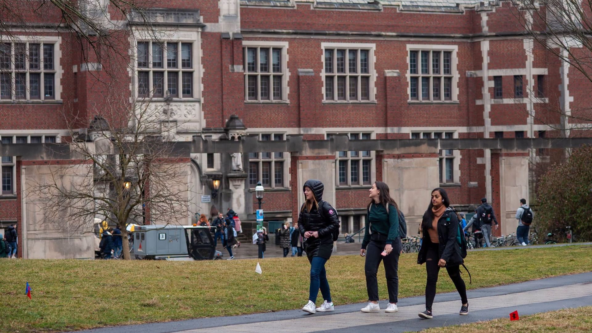 Students walk across a grassy campus path in front of a large red brick Princeton University building on an overcast day.