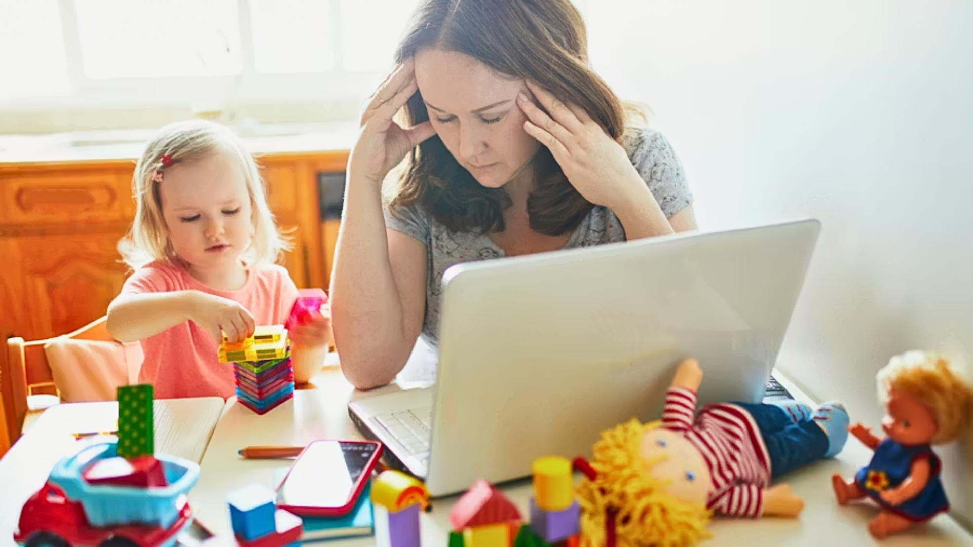 A medium shot of a stressed-looking woman with her hands to her temples, sitting at a laptop covered in toys; next to her, a young girl in a pink shirt plays with colorful building blocks, symbolizing the mental load of working from home with children.
