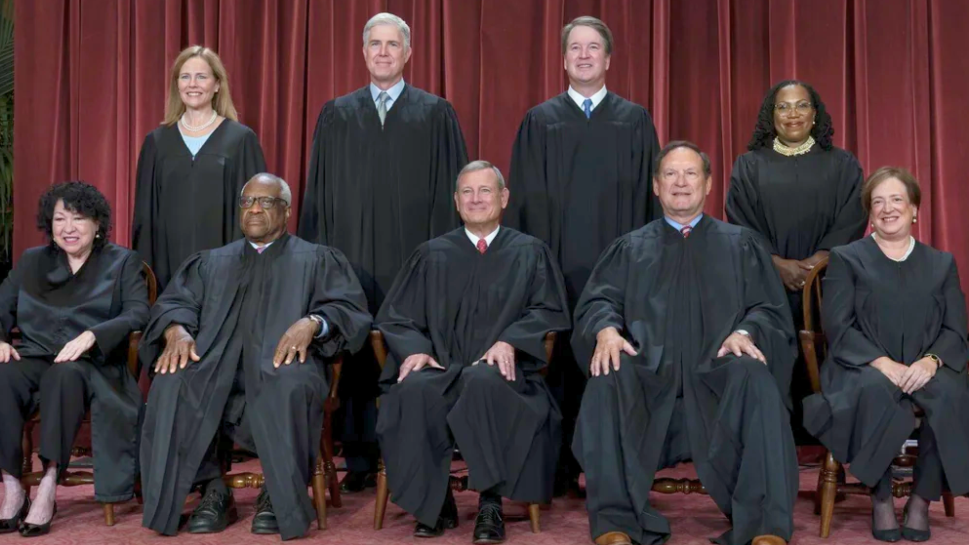 A formal group portrait of the nine Supreme Court Justices in their black judicial robes, arranged in two rows—four standing and five sitting—against a backdrop of deep red curtains.