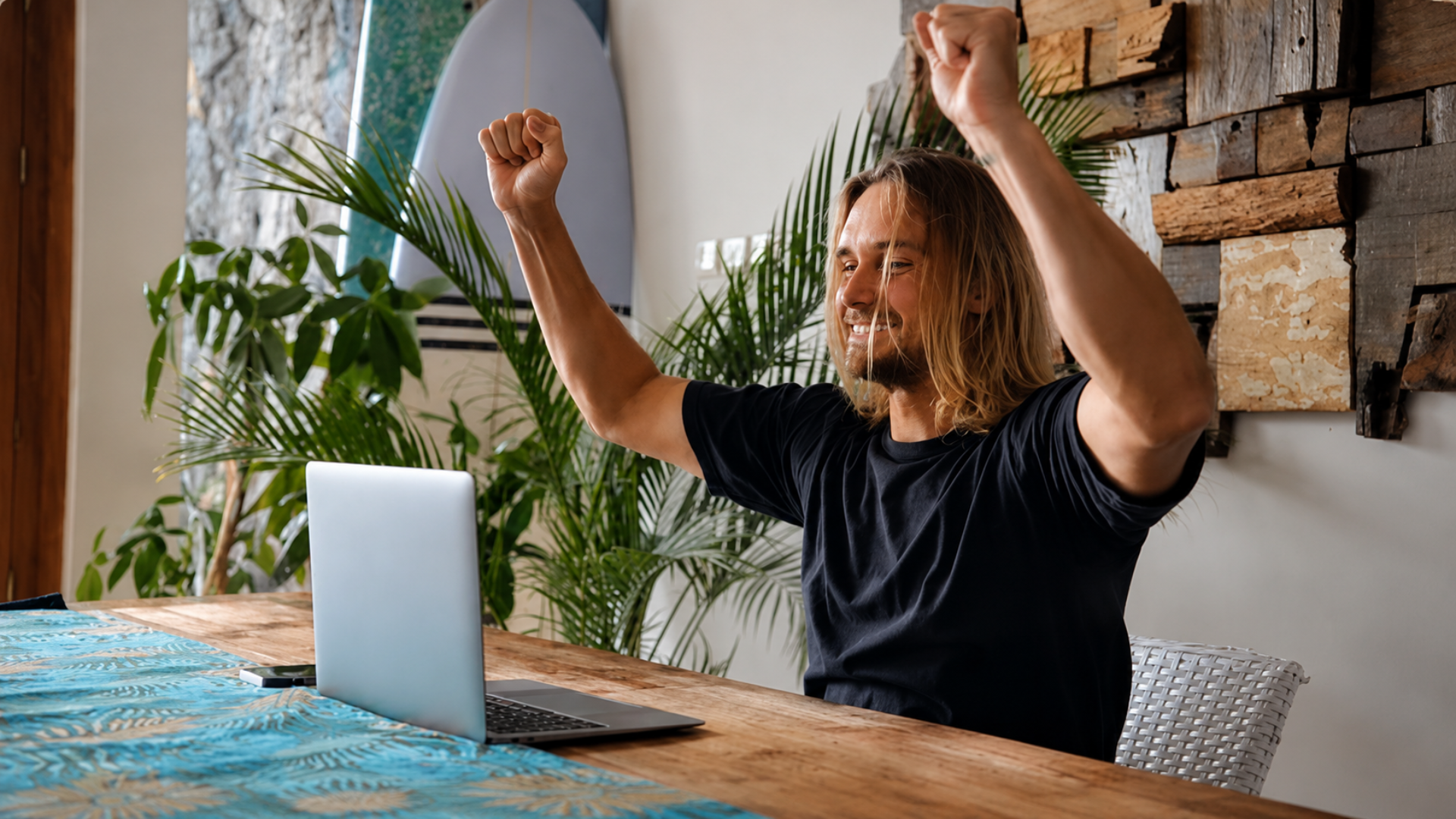 A medium shot of a man with long blond hair and a black t-shirt sitting at a wooden table, cheering with both fists raised in the air while looking at a laptop; the room has a tropical feel with large palm leaves and a surfboard in the background.