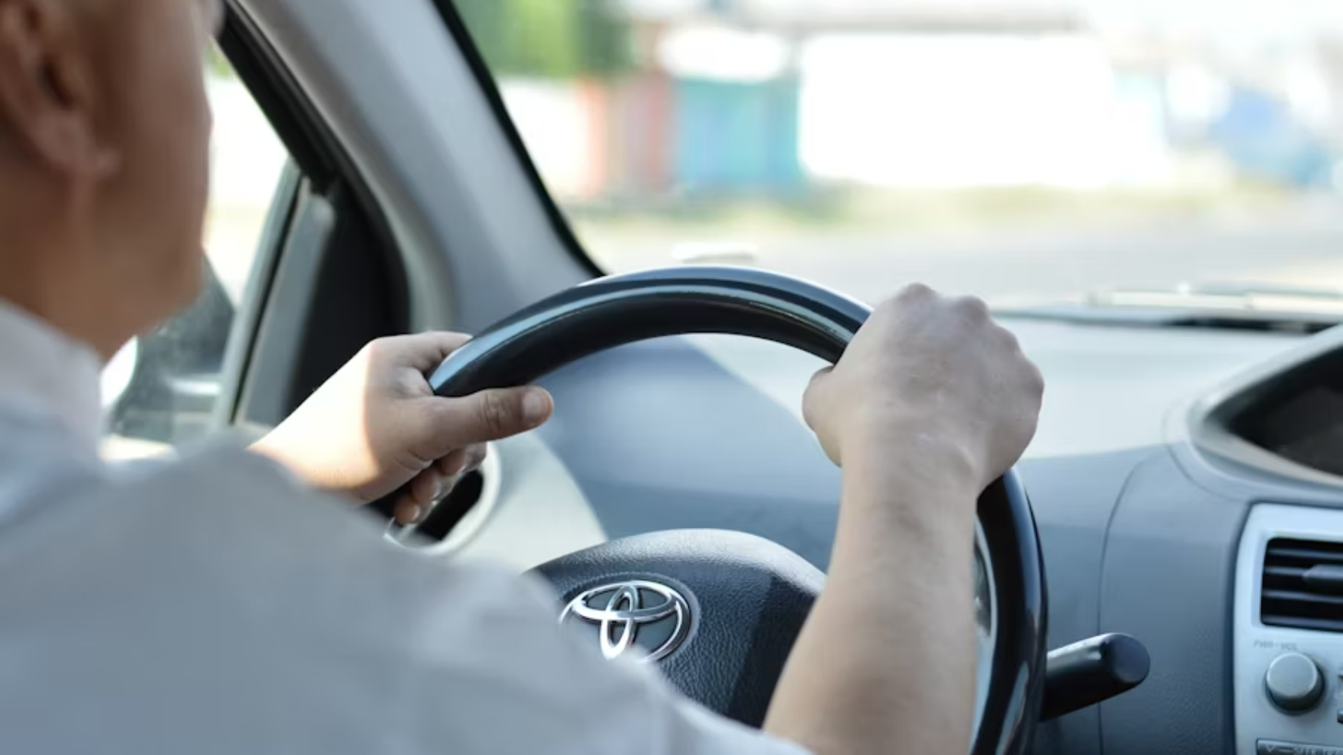 A close-up shot from the back seat of a vehicle looking forward, showing a man's hands firmly gripping a black steering wheel with a visible chrome Toyota logo in the center; the background shows a blurred, sunlit road ahead.