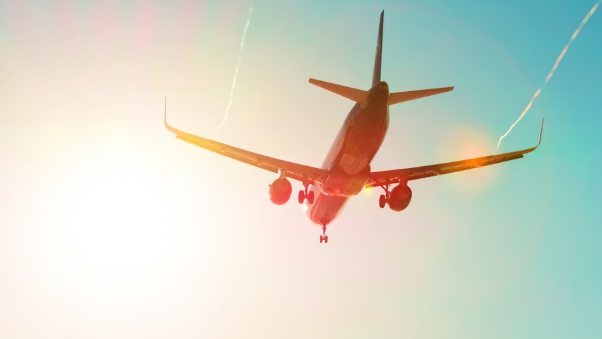 A bottom-up shot of a passenger jet with its landing gear down, silhouetted against a bright, hazy sun and a teal sky.