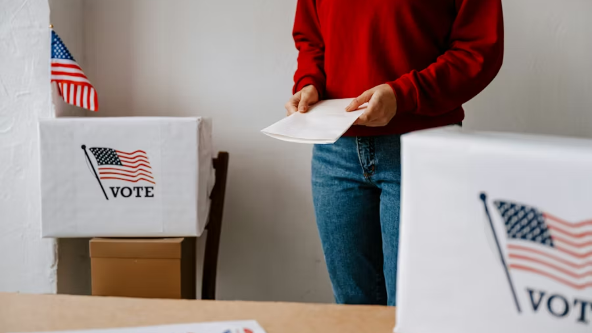 A close-up shot focusing on the midsection of a poll worker in a red sweatshirt and blue jeans holding a white paper envelope containing a ballot; the worker stands between two large, white cardboard drop boxes draped with an American flag graphic that reads "VOTE."