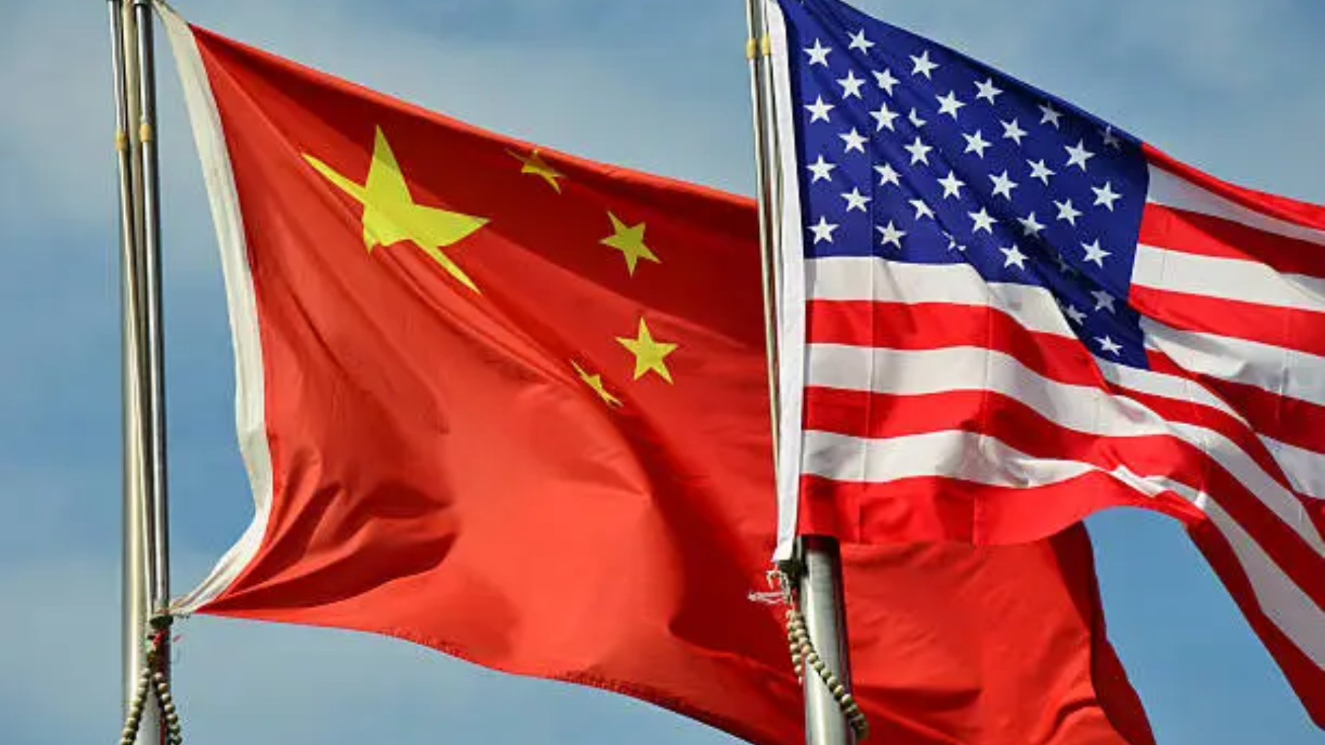 A close-up shot of the national flags of the People's Republic of China and the United States of America flying side by side on metal poles against a blue sky.